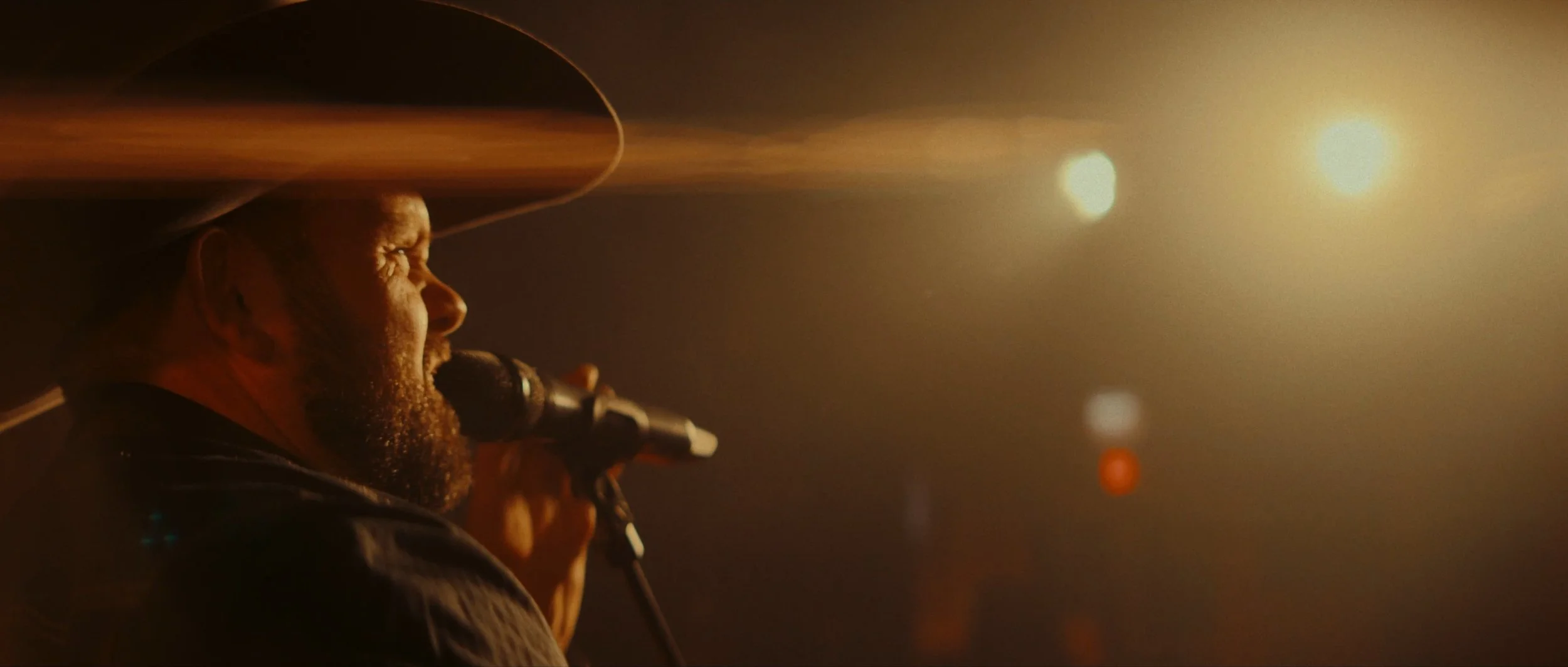 A man wearing a beige sombrero with a wide brim, singing into a microphone on stage, lit by warm, dramatic lighting with bright spots and a blurred background.