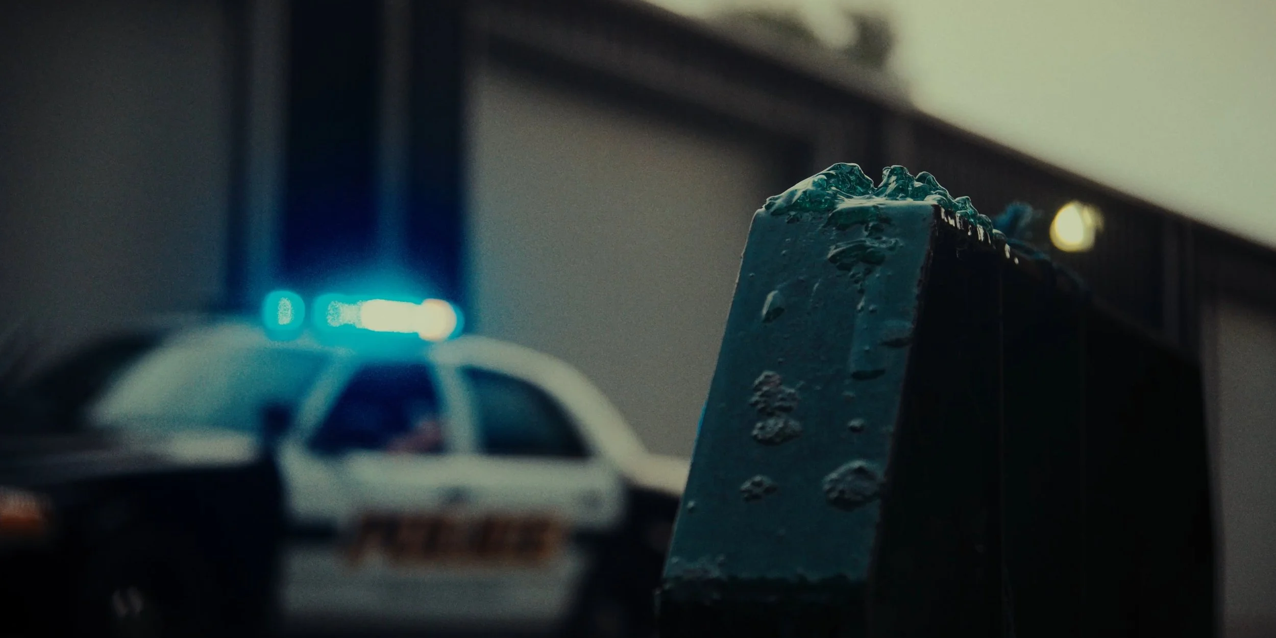 Close-up of a toy police car and a piece of broken ice on a table, with blurred background of a building and glowing blue lights.