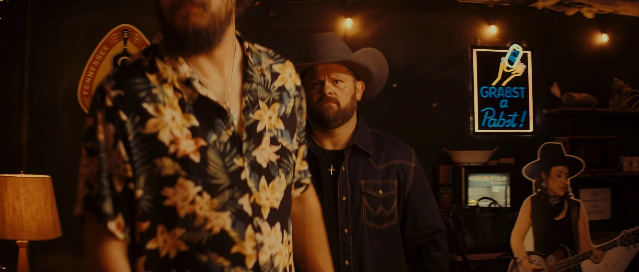 Two men in a dimly lit bar, one with a beard wearing a floral shirt, and the other with a beard and a cowboy hat, looking at each other. Behind them, a woman with a hat playing guitar is visible, and a neon sign that reads "Grabst a Pabst!" is on the