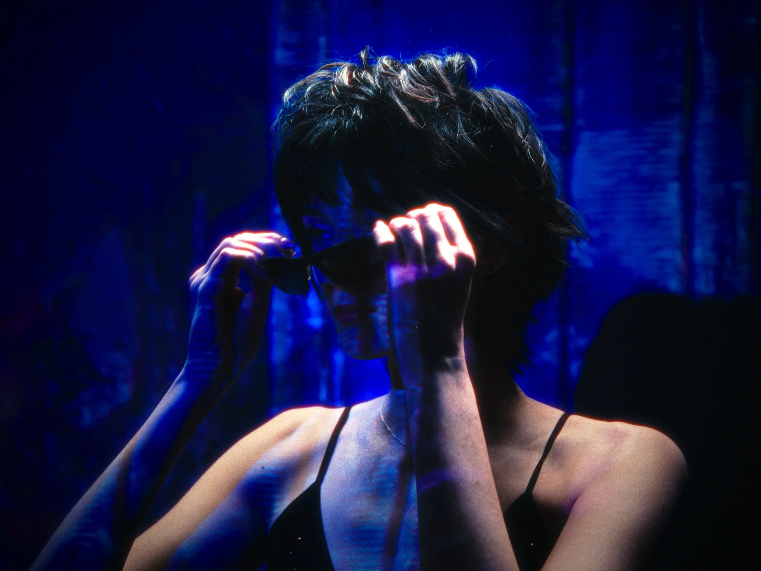 A woman with short dark hair wearing sunglasses, adjusting them with both hands, in a dark environment illuminated by blue and purple lights.