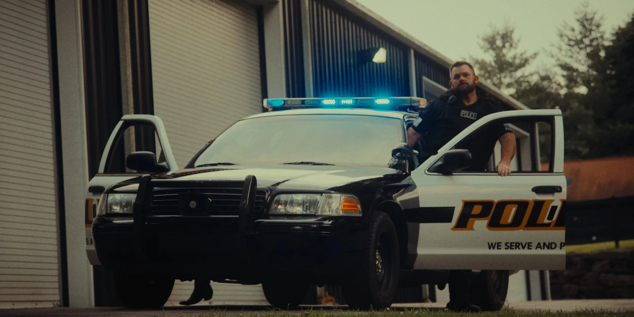 A police officer with a beard stands in the open door of a police car with blue flashing lights, parked outside a building with a metal roll-up door during daytime.