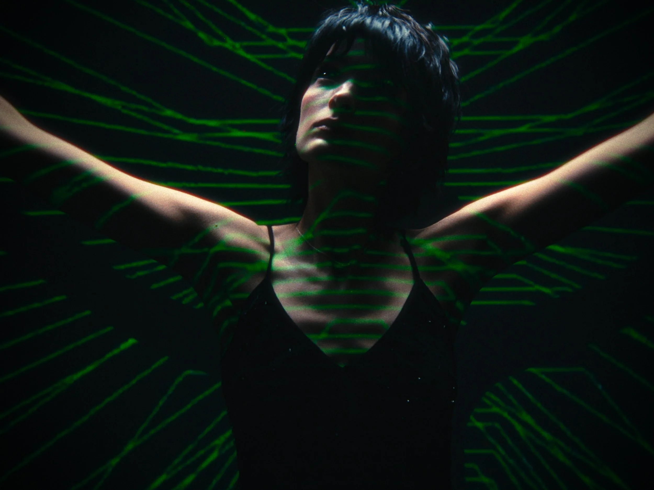 A woman with short dark hair poses with her arms raised, wearing a black top, with green laser light projections creating patterns across her face and body, set against a dark background.