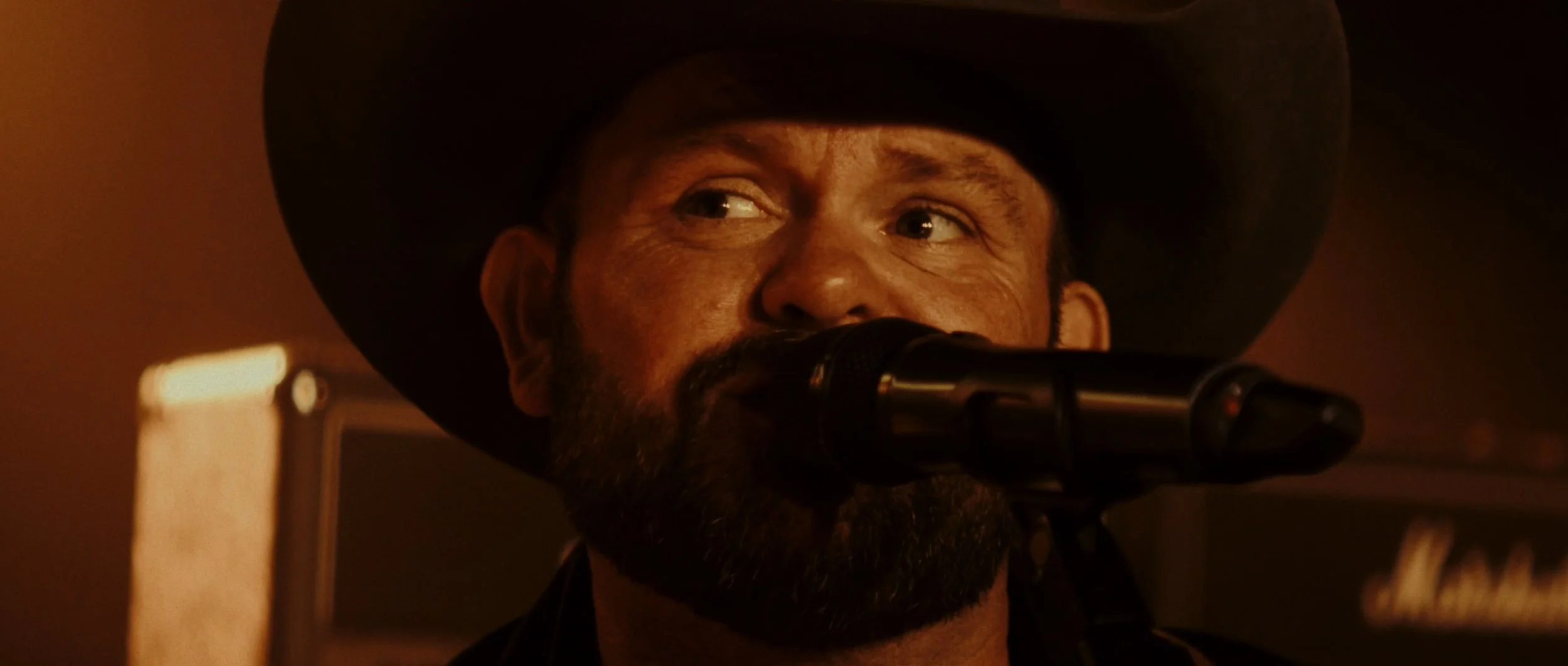 A man with a beard wearing a black cowboy hat, speaking or singing into a microphone in a dimly lit room.