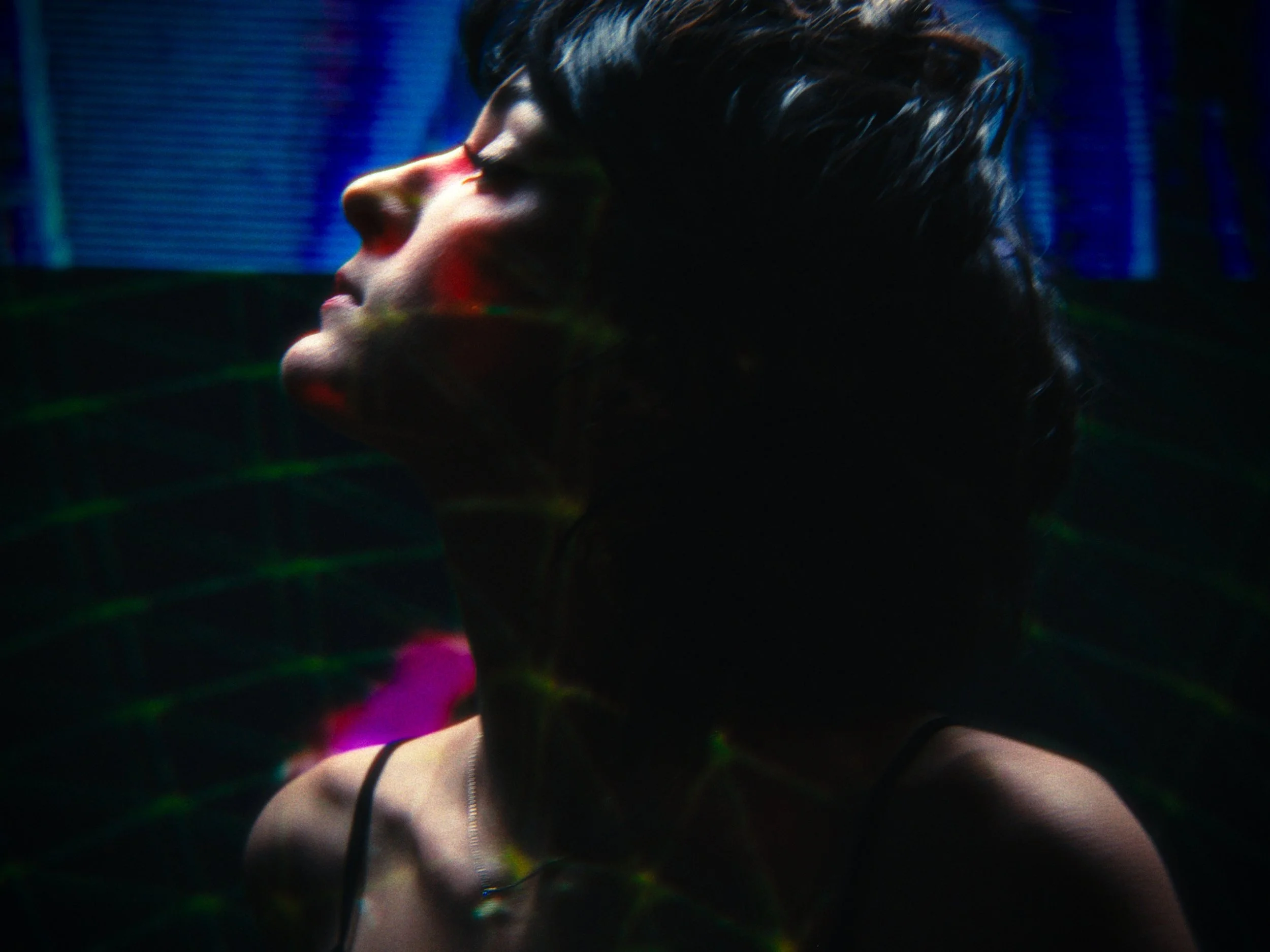 A person with short dark hair and light skin appears to be in a dark, neon-lit environment, possibly a nightclub or concert. Their face is turned to the side with eyes closed, and they are wearing a thin strap top.