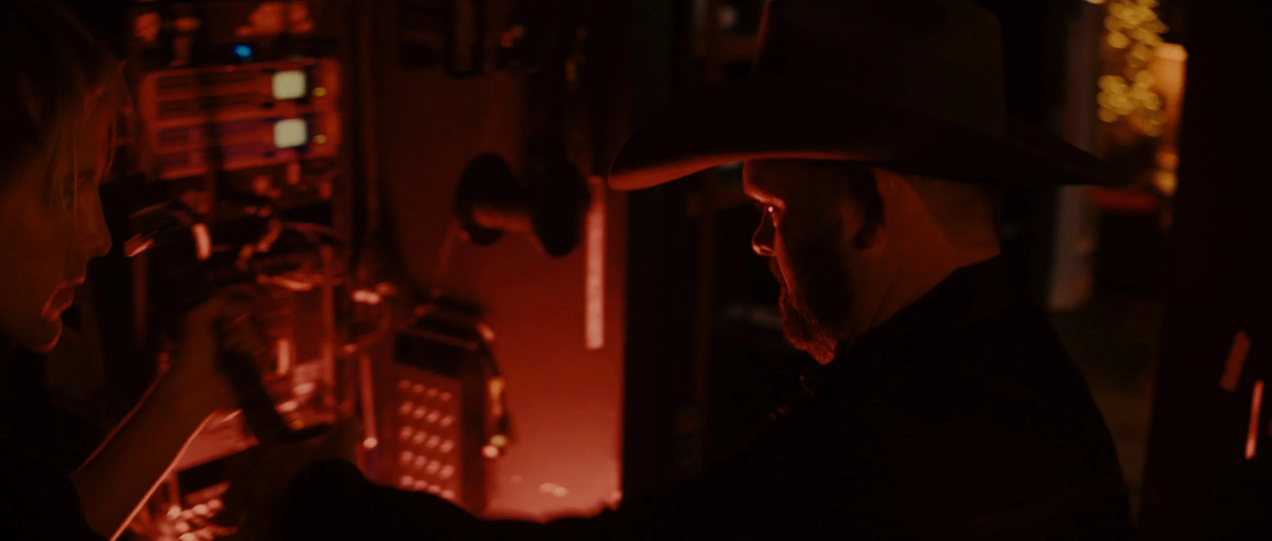 A man wearing a wide-brimmed hat and glasses working on audio equipment in a dark environment with red lighting.