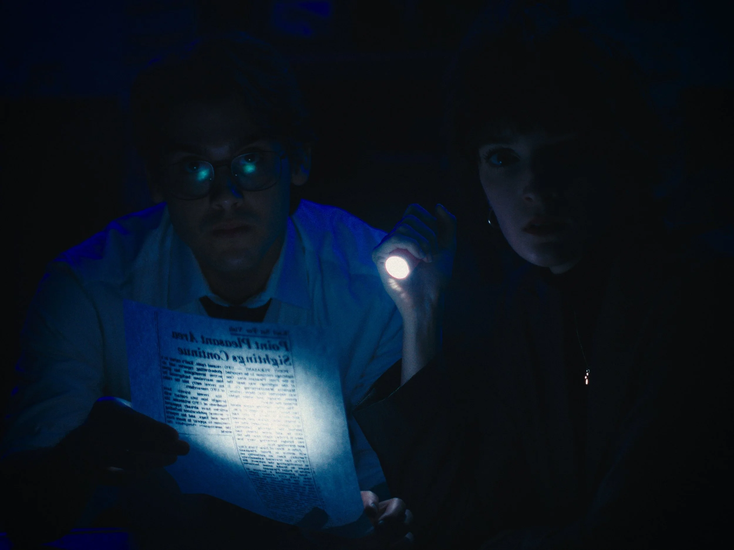 Two people in a dark room, one holding a flashlight and the other holding a newspaper.