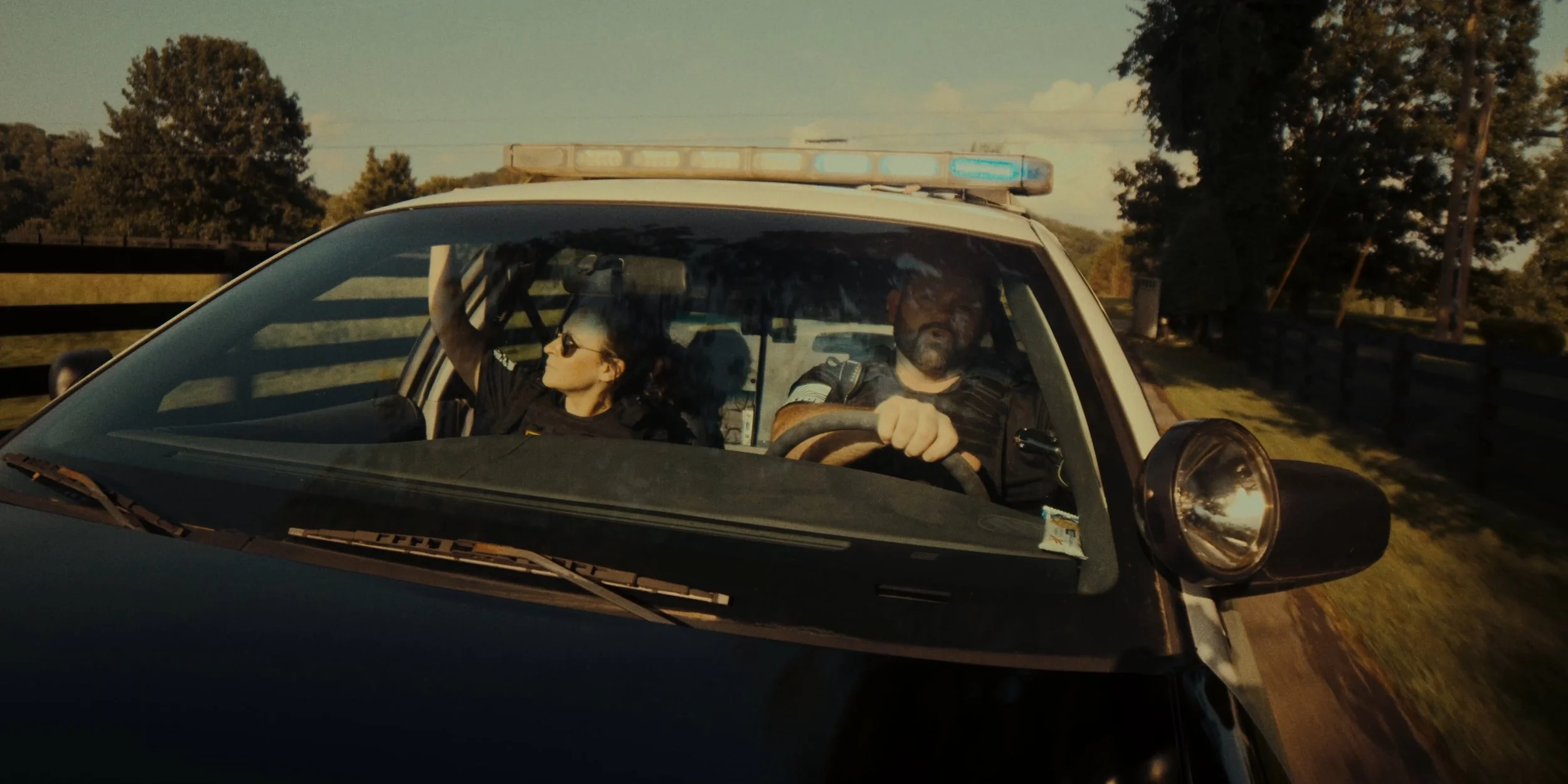 A police car with two officers inside, one driving and the other sitting in the passenger seat, is on a rural road during sunlight, with trees and a fence in the background.