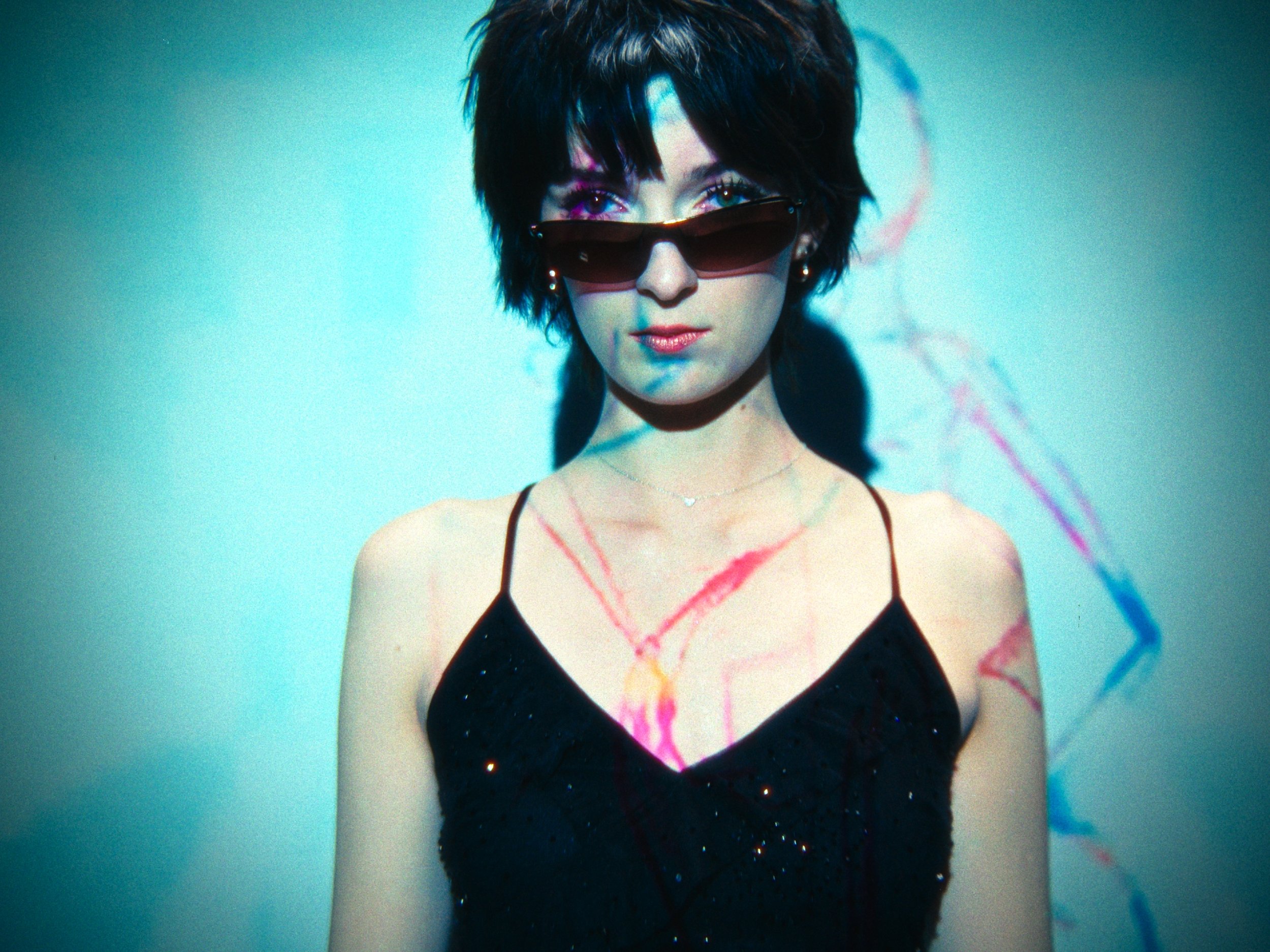 A woman with short dark hair wearing sunglasses, a black spaghetti strap dress, and a delicate necklace, standing against a plain background with abstract colorful lines.