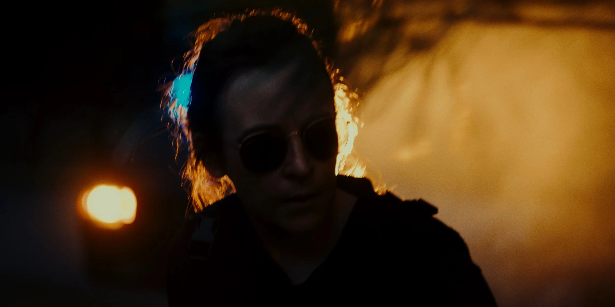 A person wearing dark sunglasses and a dark jacket is illuminated by a fiery orange background with smoke and bright lights, creating a dramatic silhouette.