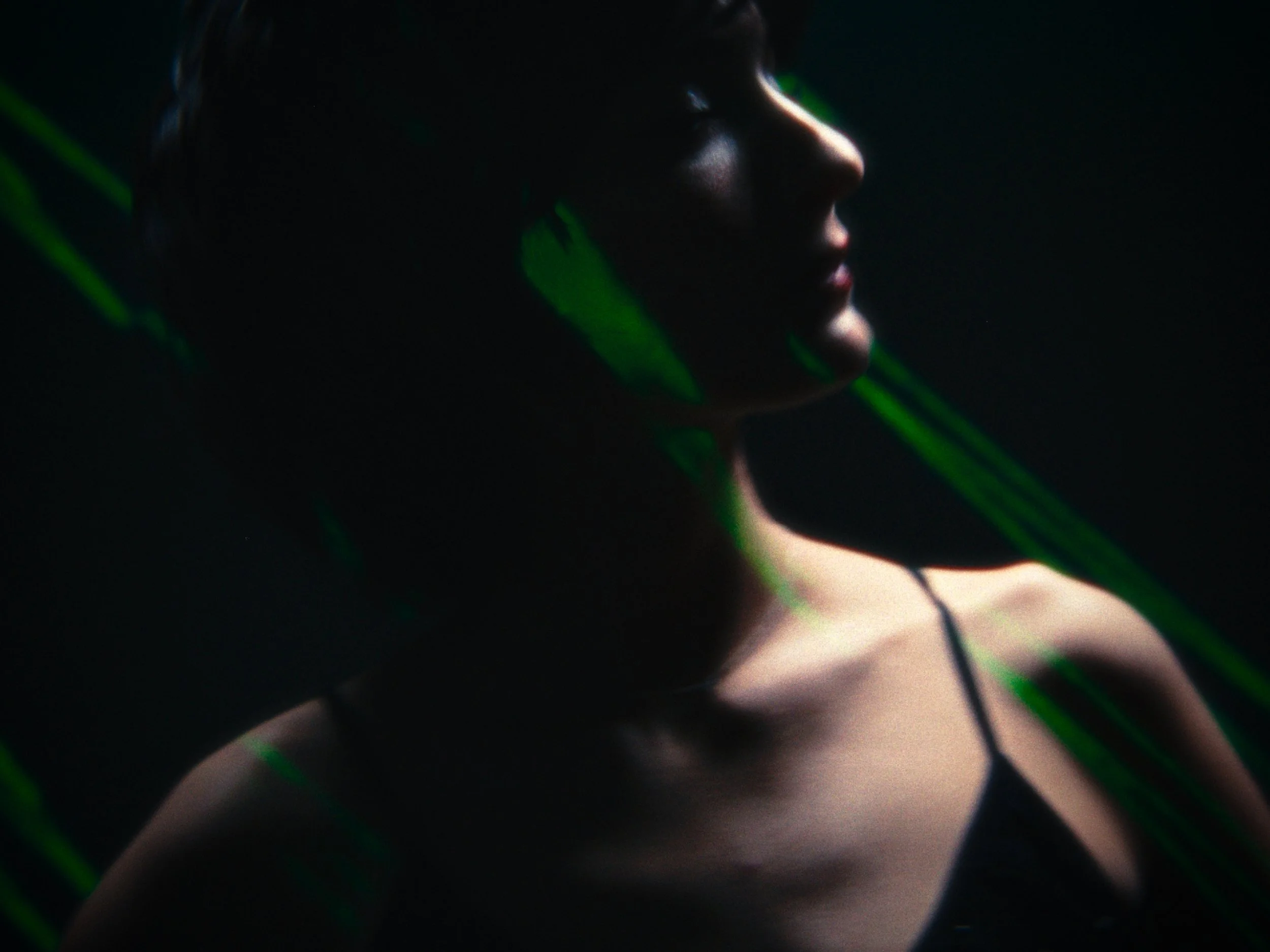 A side profile of a woman's face and shoulders illuminated softly in a dark setting with green light streaks across her face and shoulders.