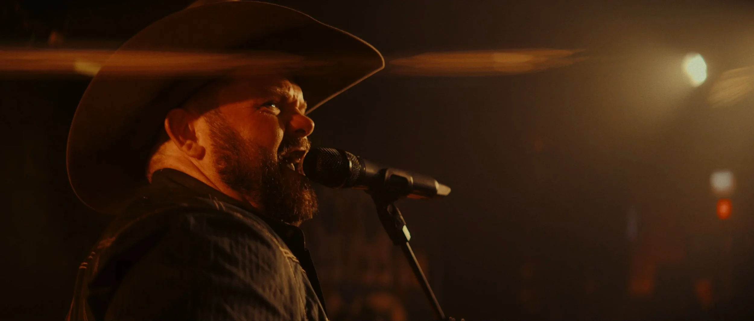 Man wearing a cowboy hat singing into a microphone with warm stage lighting.