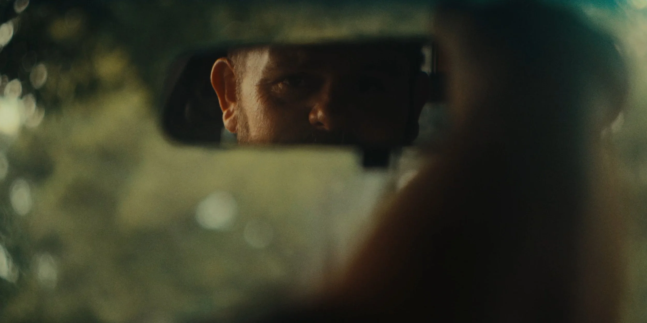 A person's face is partially visible through a car's rearview mirror, with their eyes and part of their face illuminated.