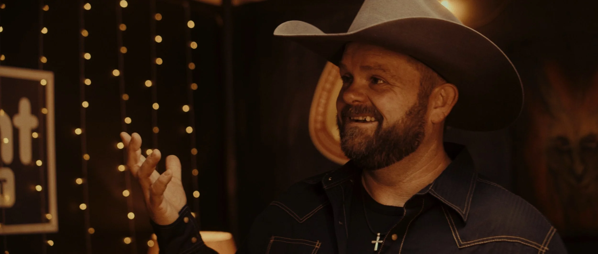 A man with a beard wearing a cowboy hat and a dark shirt with stitching, smiling and gesturing with his right hand in a warmly lit room decorated with string lights and western-themed artwork.