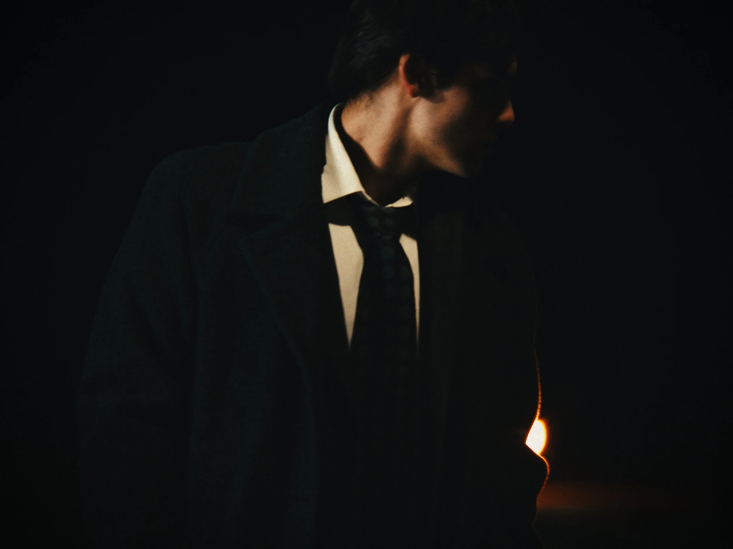 A man dressed in formal attire, including a dark coat, white shirt, and black tie, standing in a dark setting with a faint light source behind him.