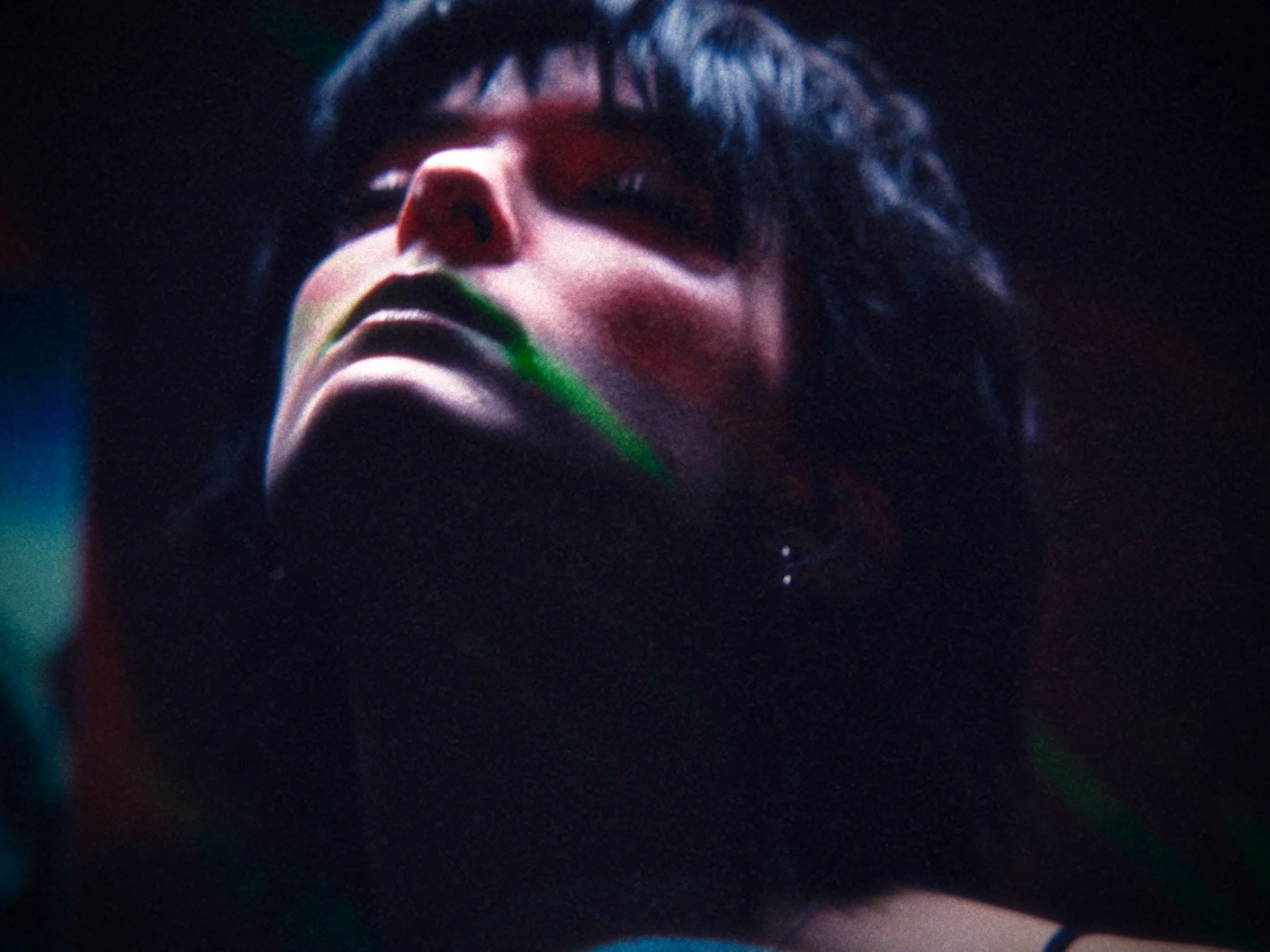 A close-up of a person's face illuminated by colorful lights, with dark hair and makeup.