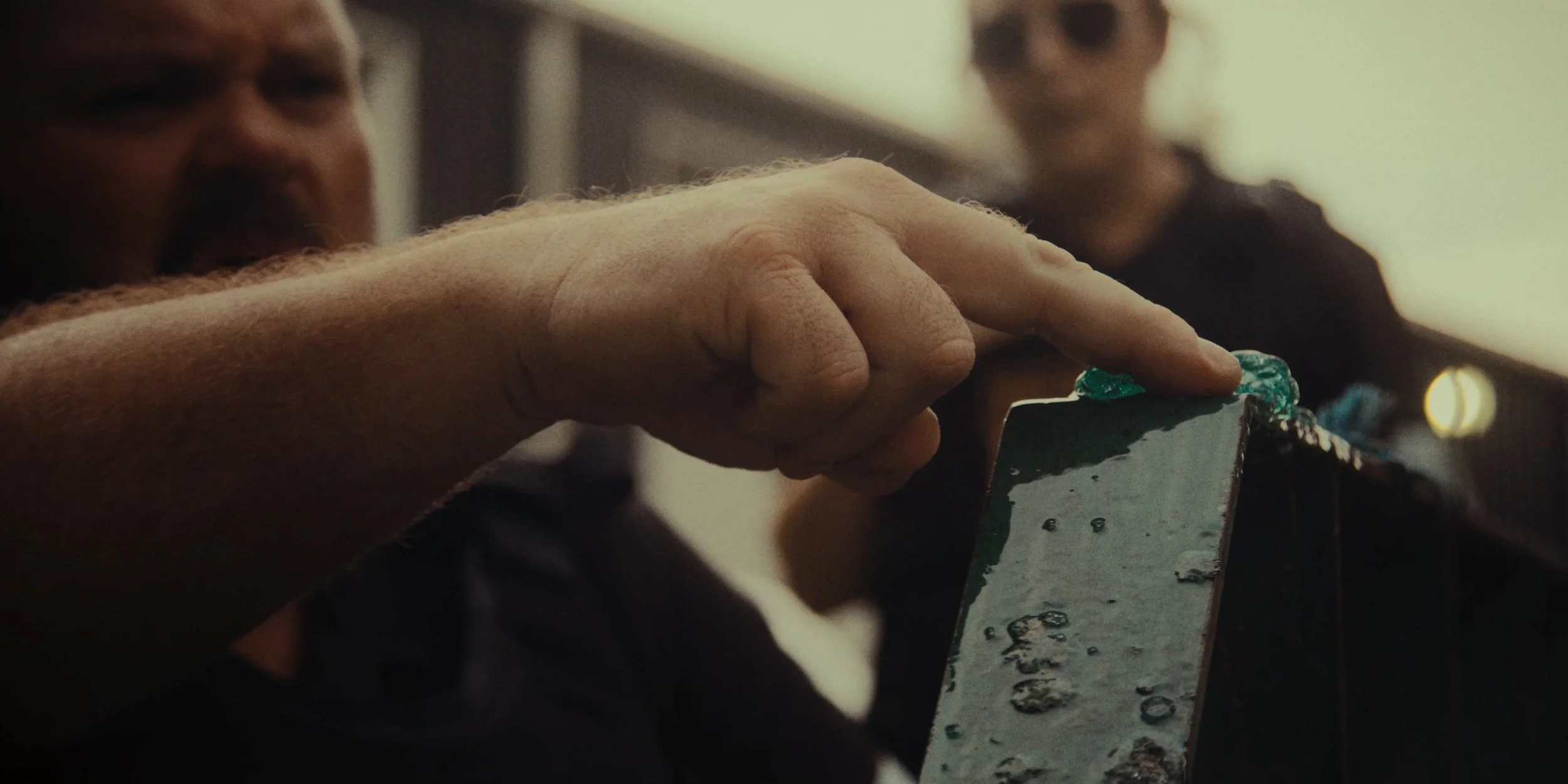 A person with a beard is pointing at a small, rectangular object with blue-green substance on top, with another person in the background.