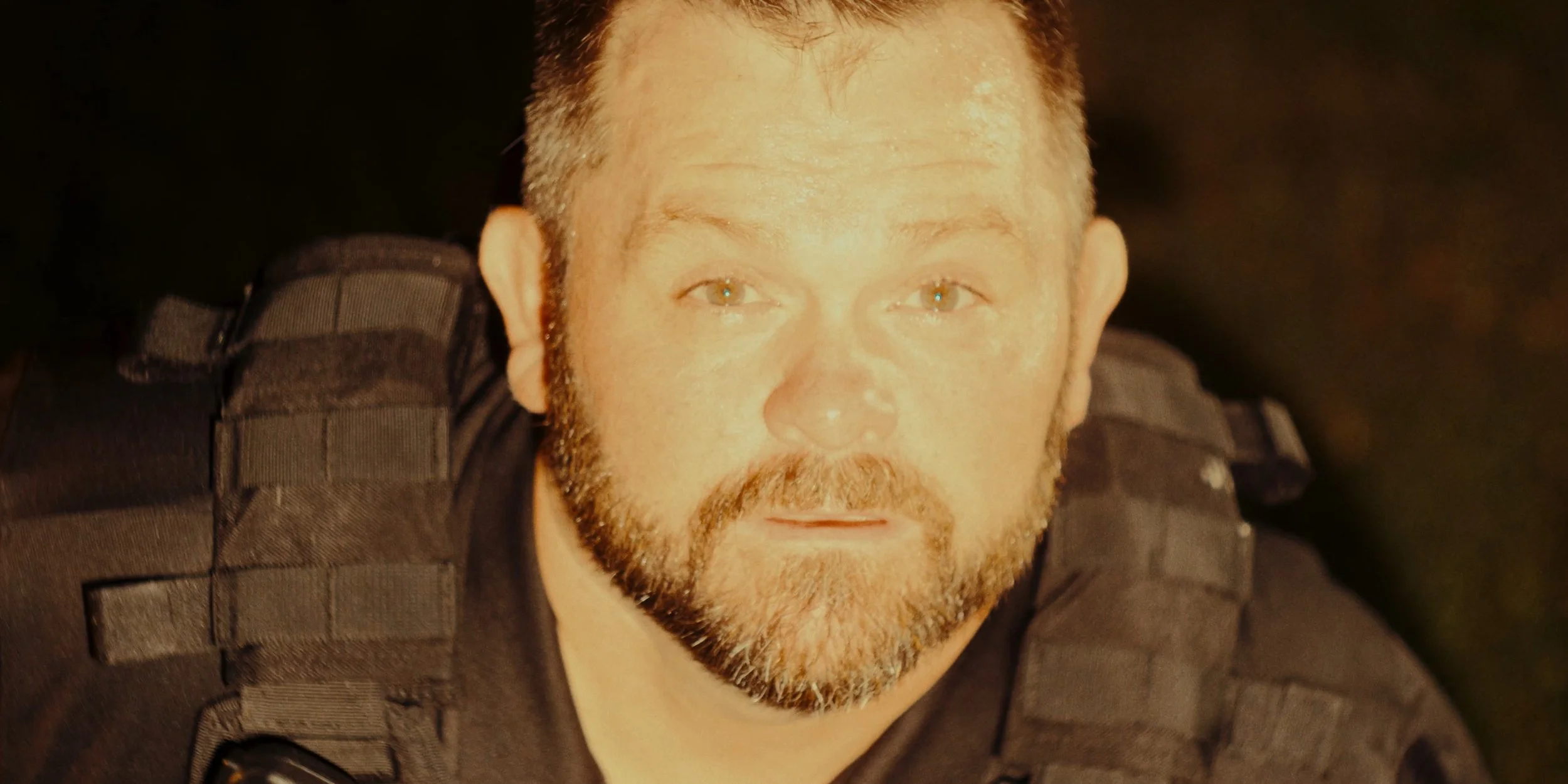 Close-up of a man with a beard in a black tactical uniform with a vest, looking directly at the camera.