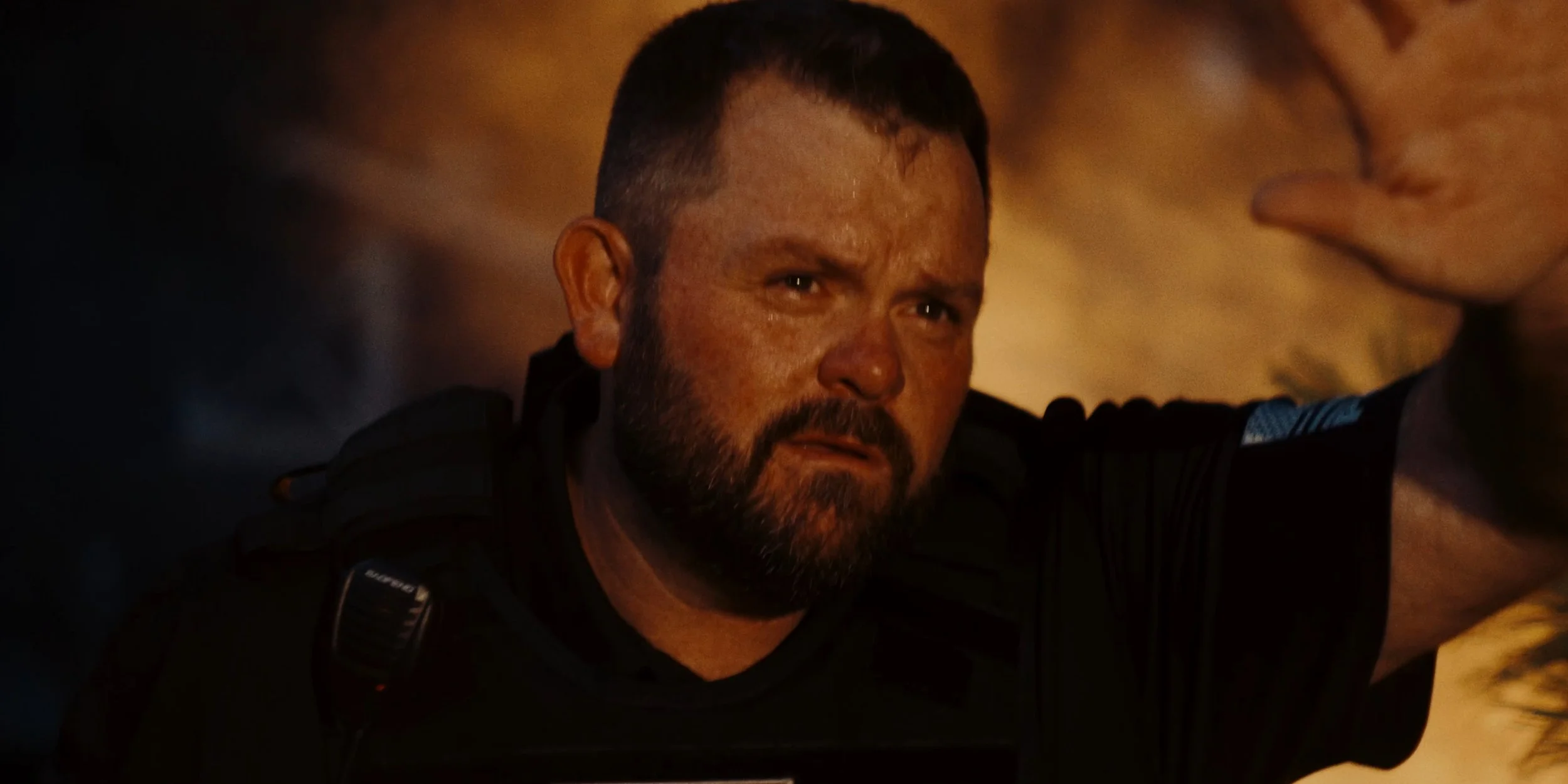 A man with a beard, short hair, wearing a black shirt and tactical gear, appears distressed, with a background that is blurred and dark.