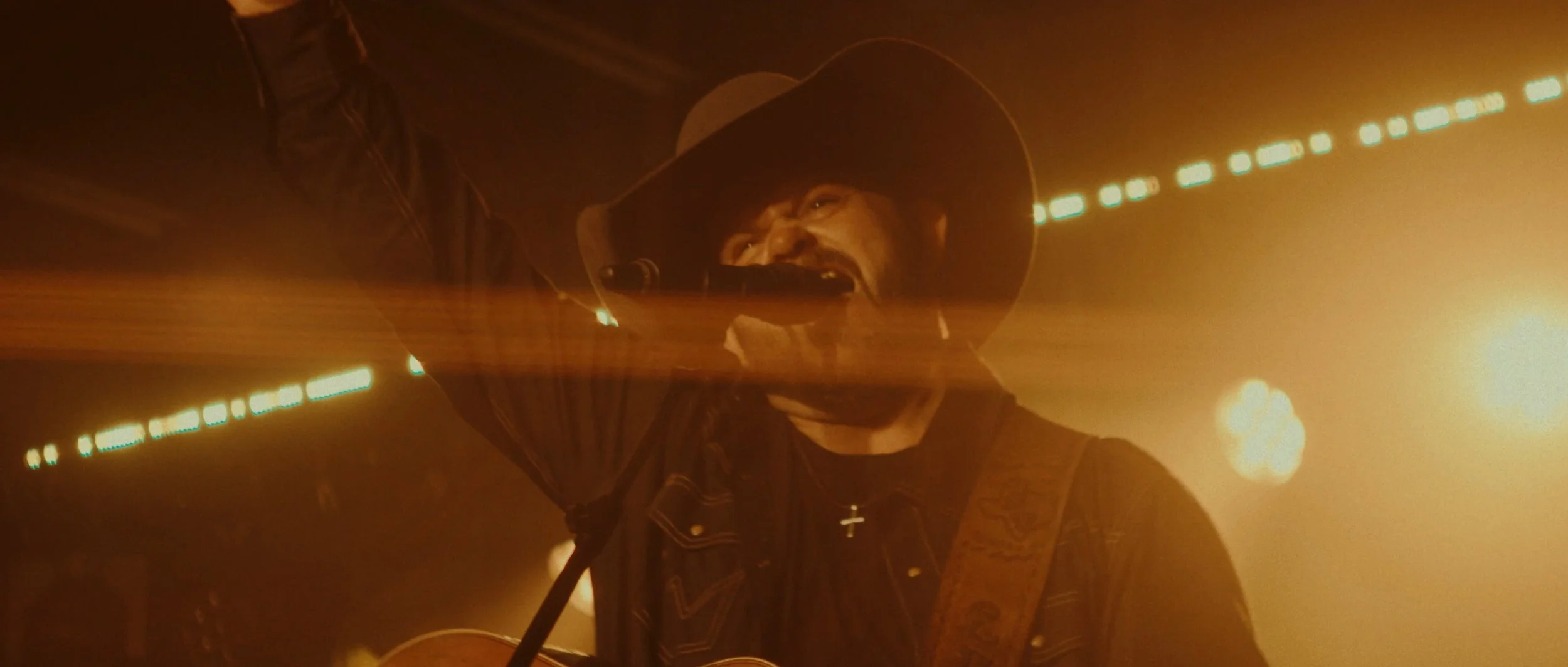 A man wearing a cowboy hat singing into a microphone on stage, illuminated by warm yellow and orange lights.