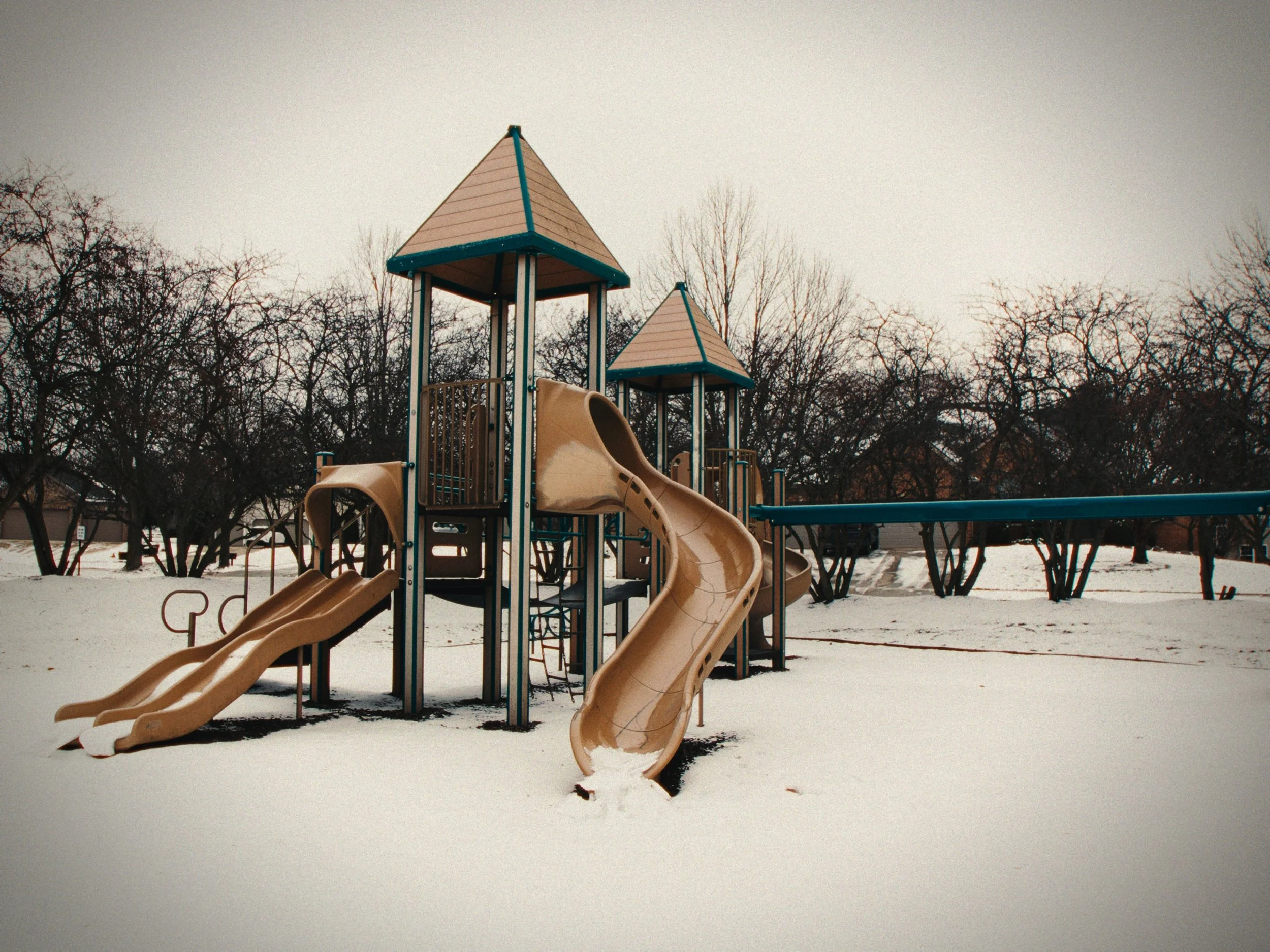 A playground with plastic slides and climbing structures covered in snow, with leafless trees in the background on an overcast day.