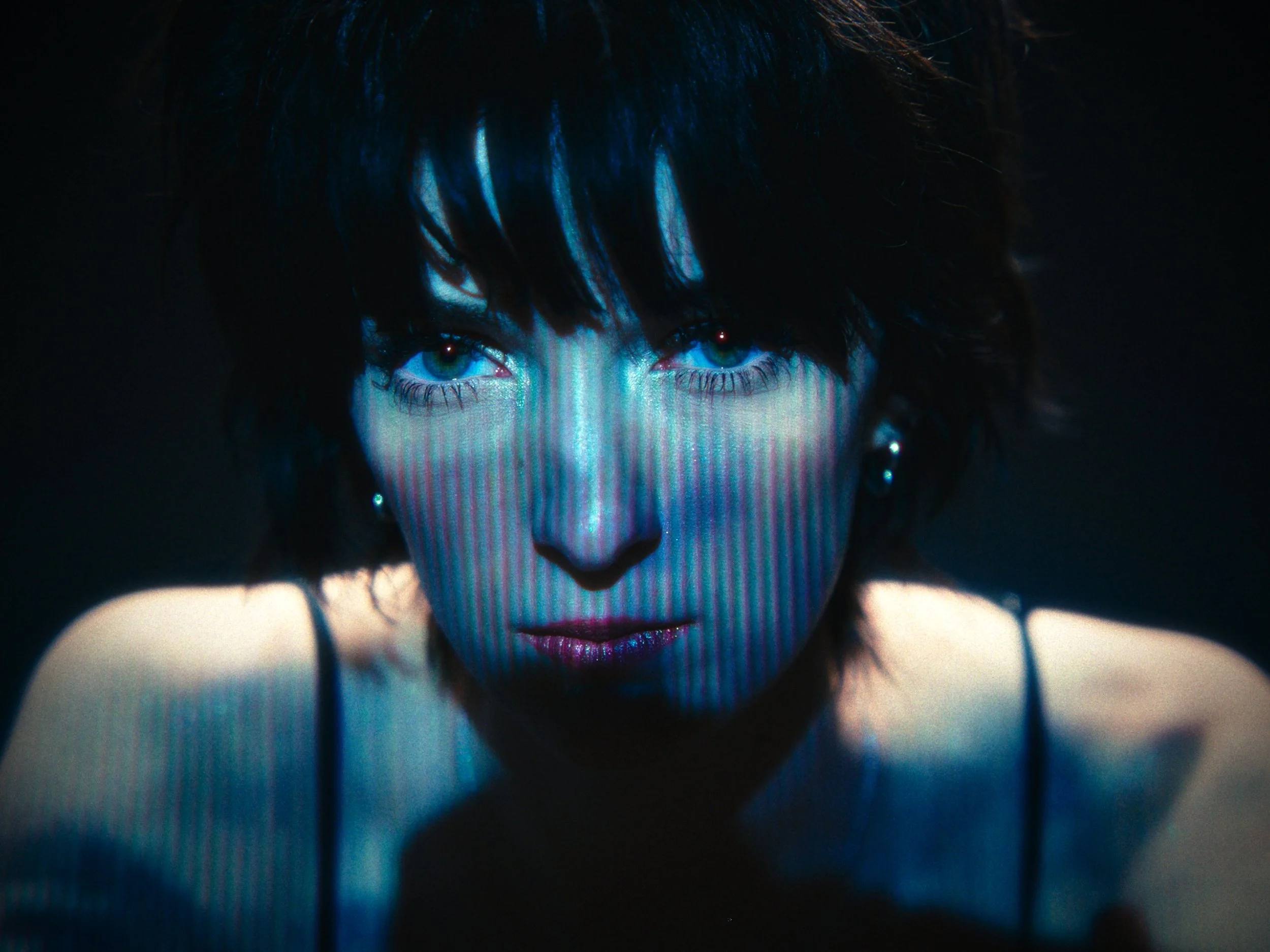 A woman with dark, short, messy hair and blue eyes looking directly at the camera. The lighting creates vertical blue and purple streaks on her face, and she is wearing a black spaghetti strap top.