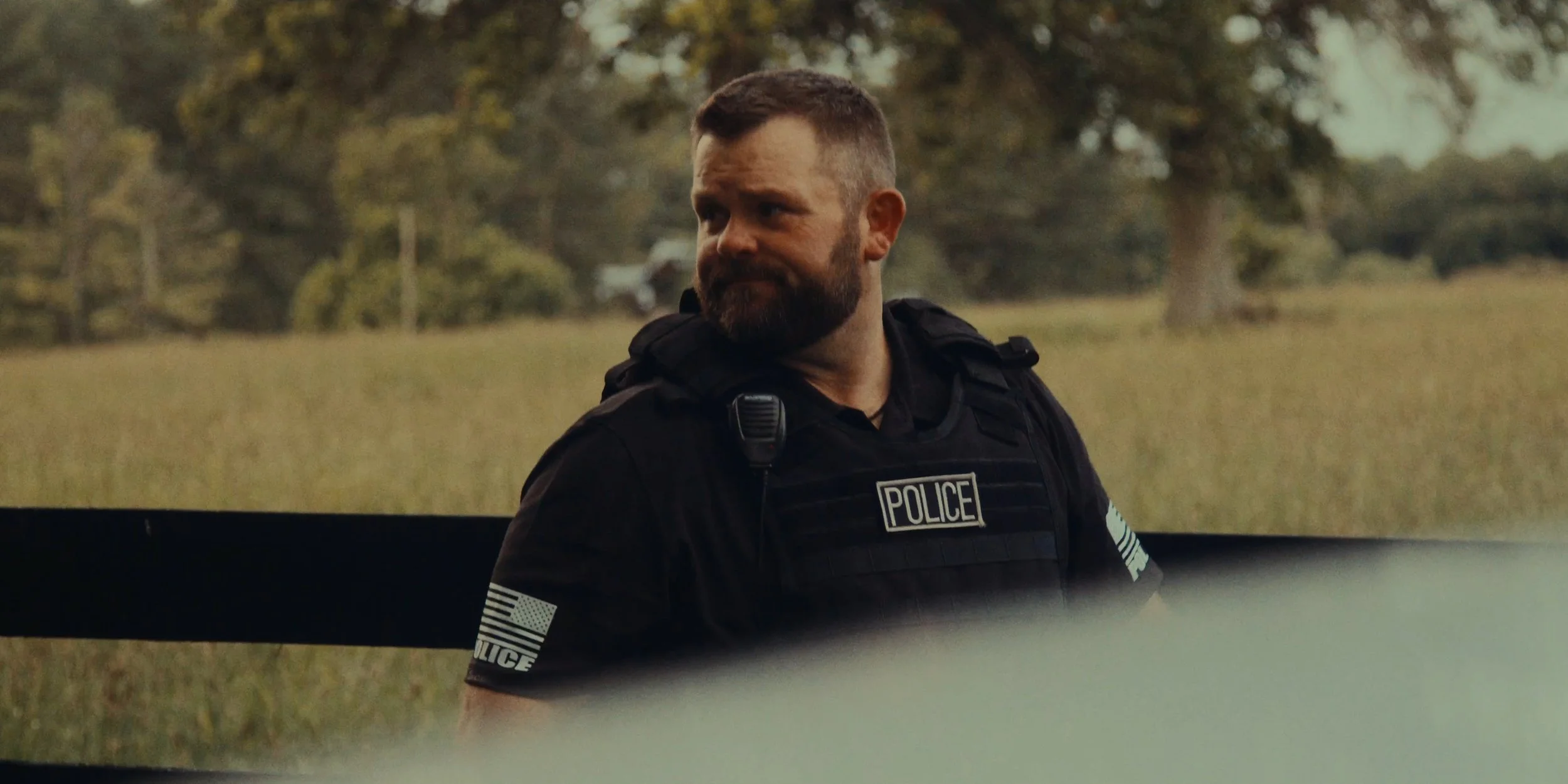 A police officer with a beard and short hair standing outdoors, wearing a black uniform with police patches on the sleeves and a badge on the chest.