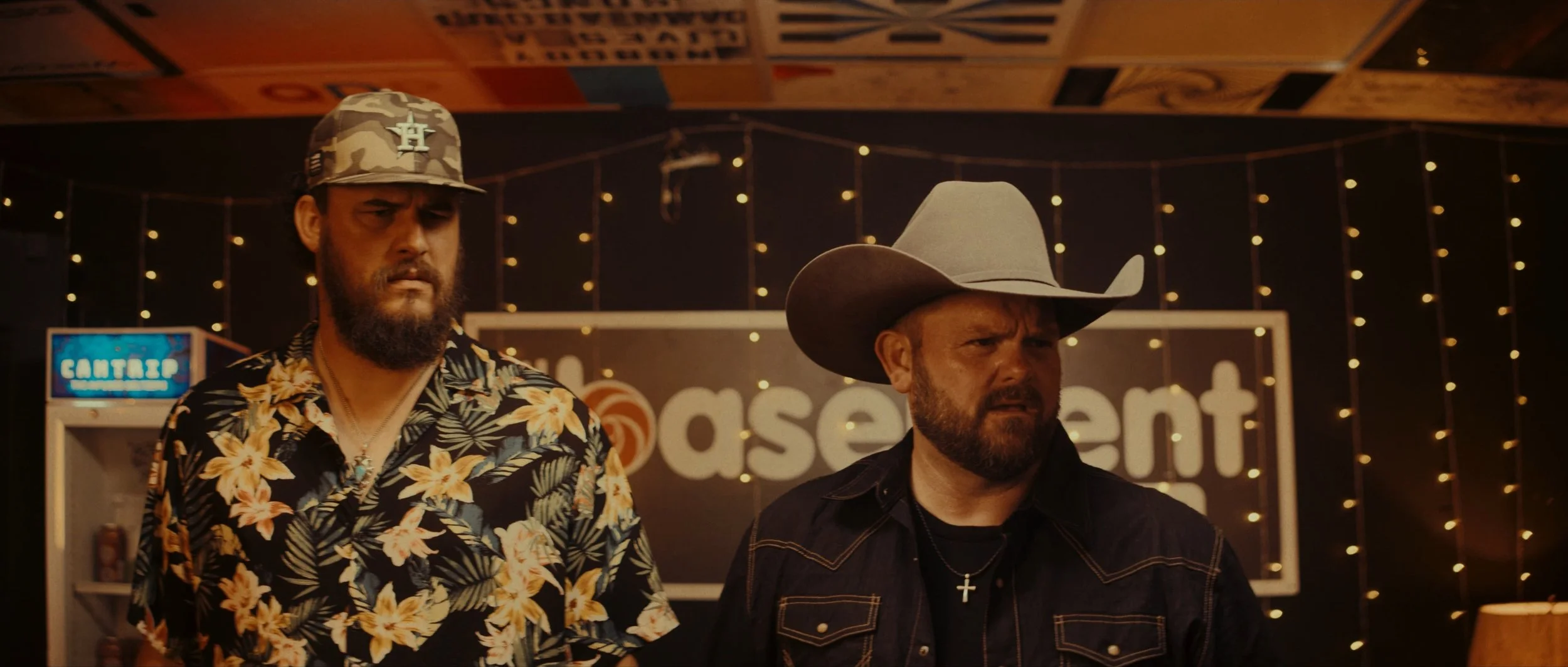 Two men with serious looks in a dimly lit room with string lights. One wears a camouflage cap and a floral shirt; the other wears a cowboy hat and a necklace with a cross. There is a large sign with the word 'basement' in the background.