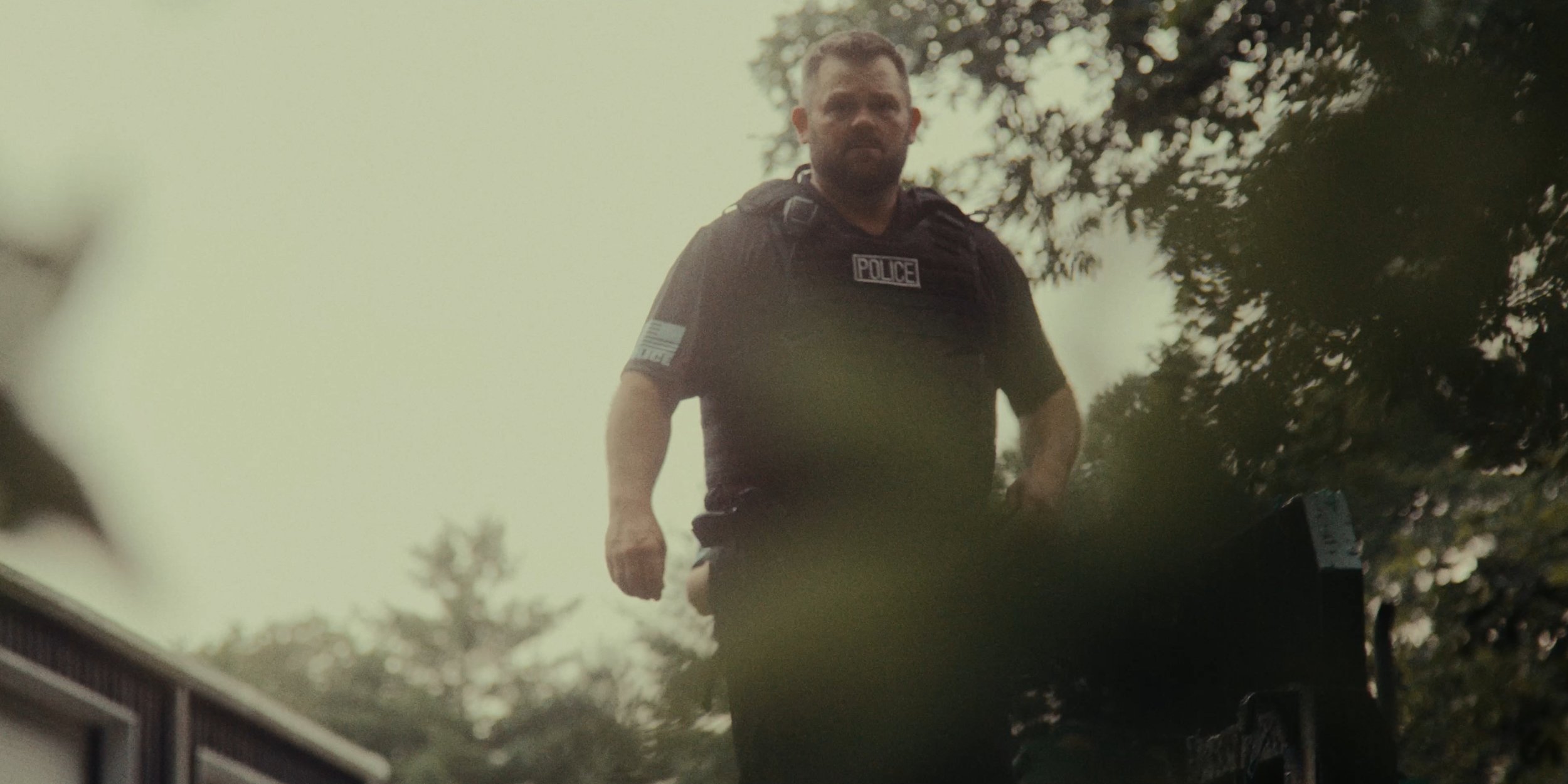 A police officer in uniform with a badge that says 'POLICE' standing outdoors near trees.