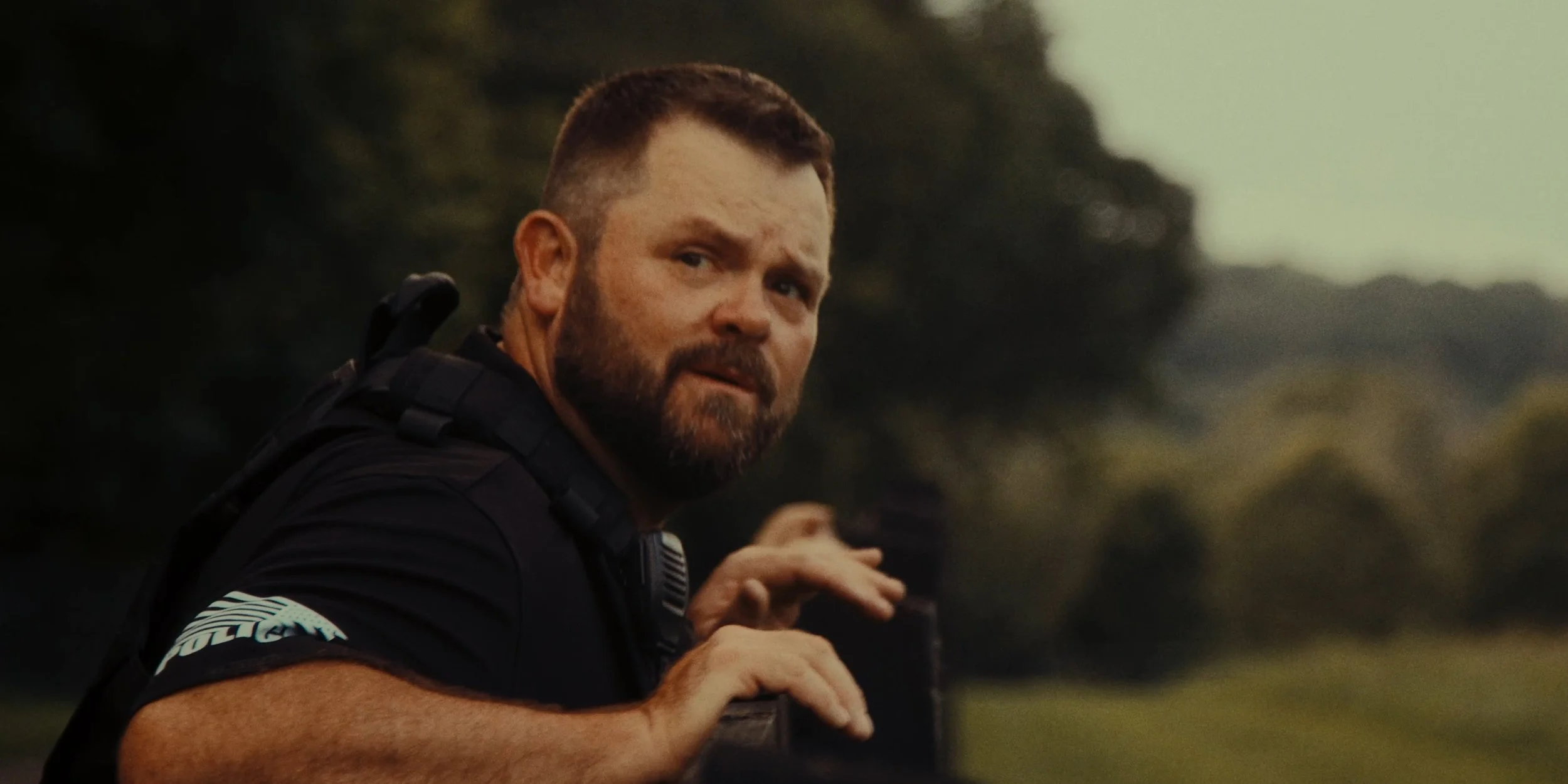 A man with a beard and short hair, wearing a black shirt with white stripes on the sleeve, looking back with a cautious expression, outdoors in a natural setting with trees and grass in the background.