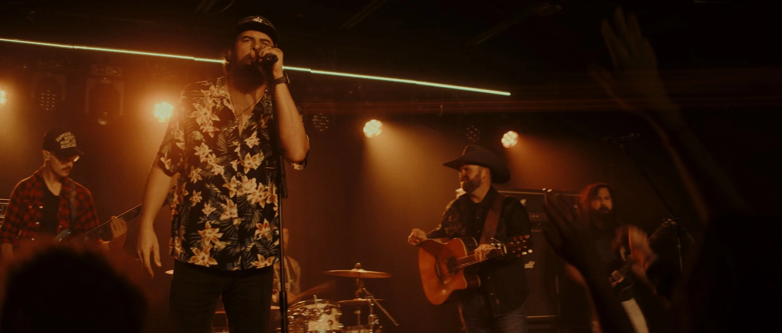 A band performing live on stage with a singer in a floral shirt, a guitarist in a cowboy hat, and other band members playing instruments in warm stage lighting.