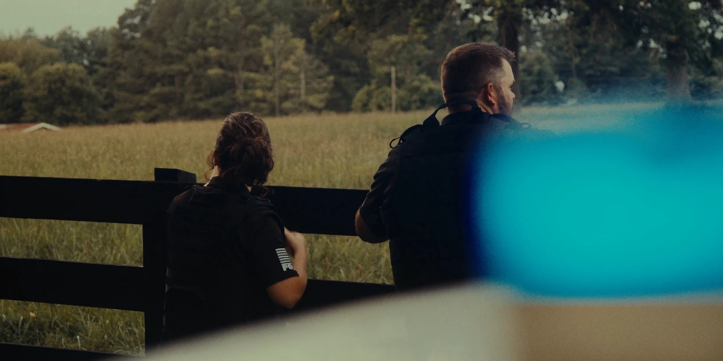 Two police officers, one woman and one man, sitting on a wooden fence, overlooking a grassy field and trees in the distance, during what appears to be late afternoon or evening.