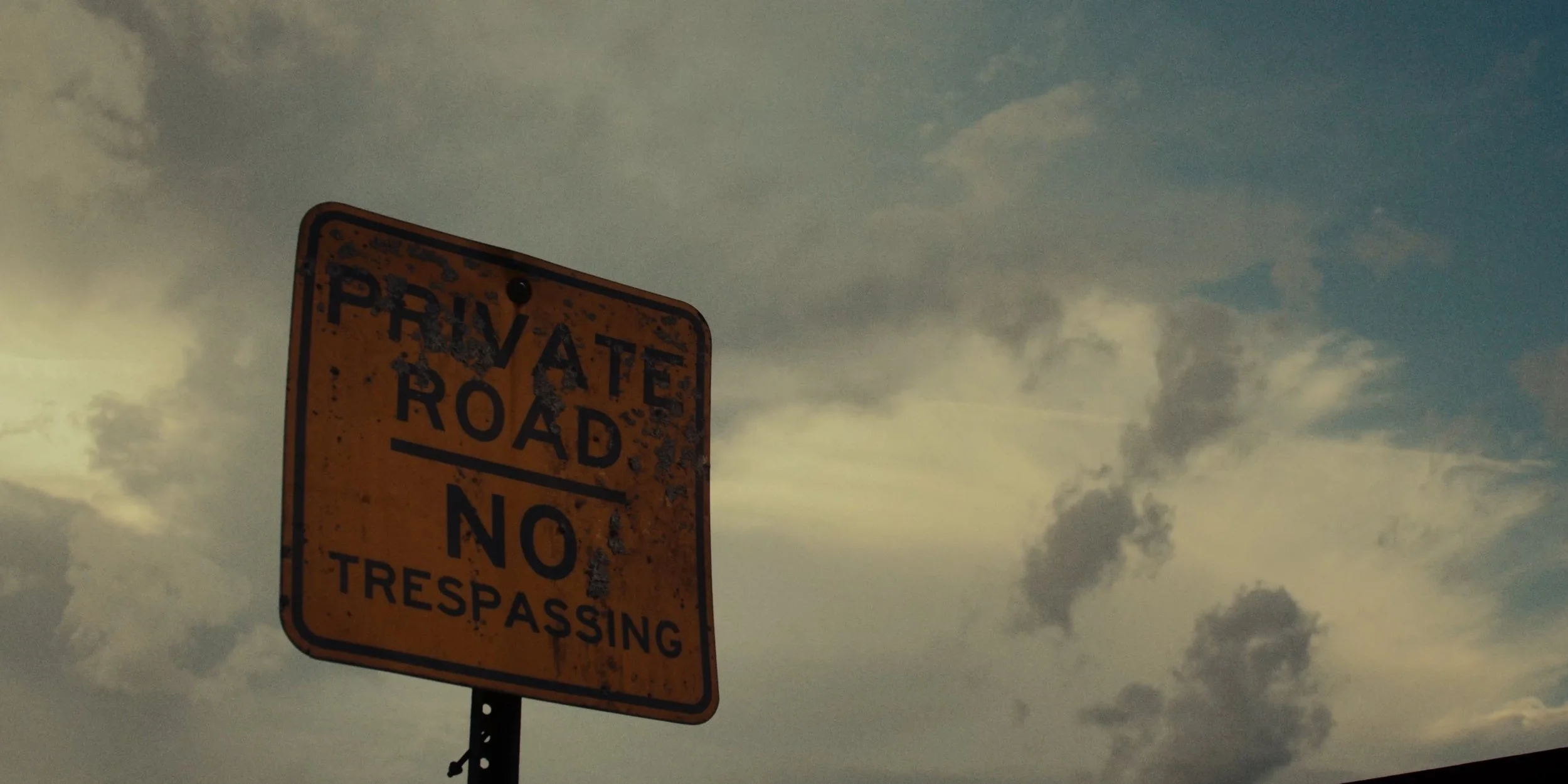 Rusty orange sign with black text reading "PRIVATE ROAD NO TRESPASSING" against a cloudy sky.