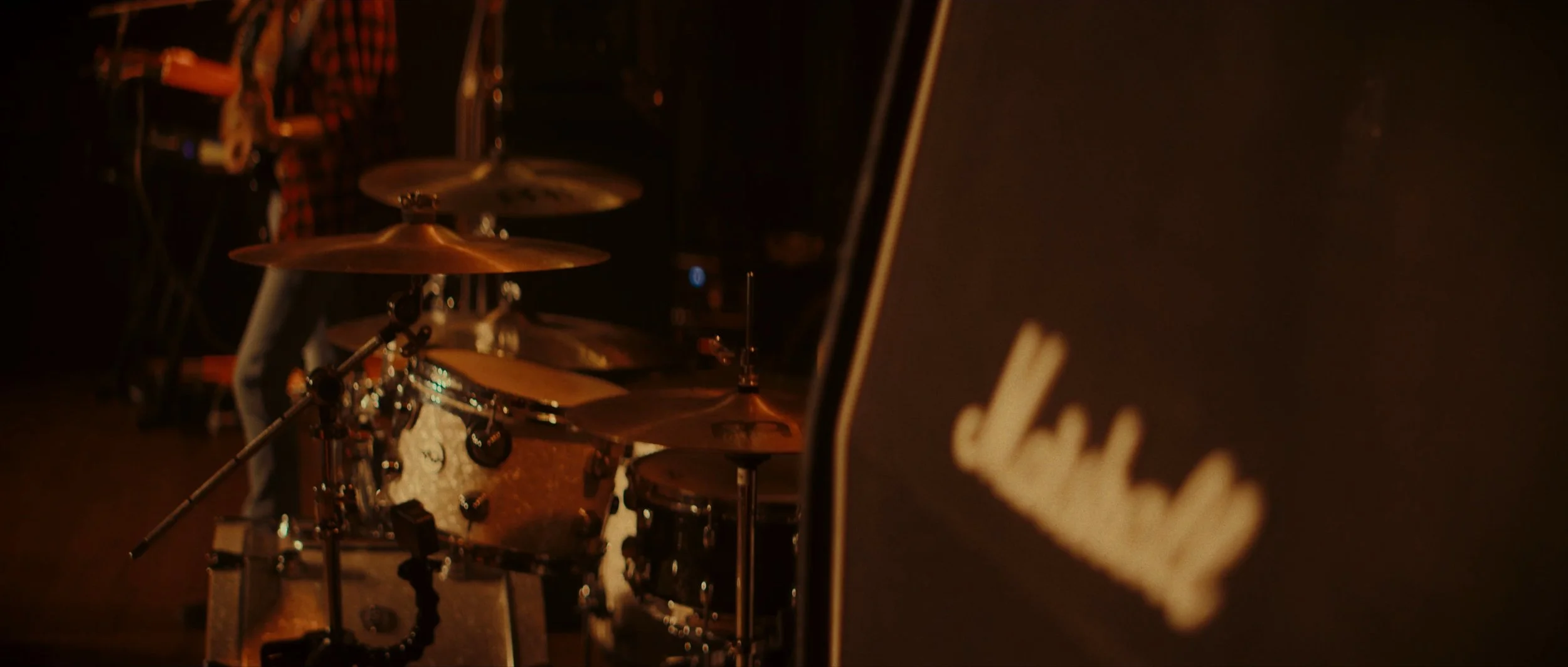 Close-up of a drum set with cymbals and drums in a dimly lit setting, with a black Marshall amplifier visible on the right.