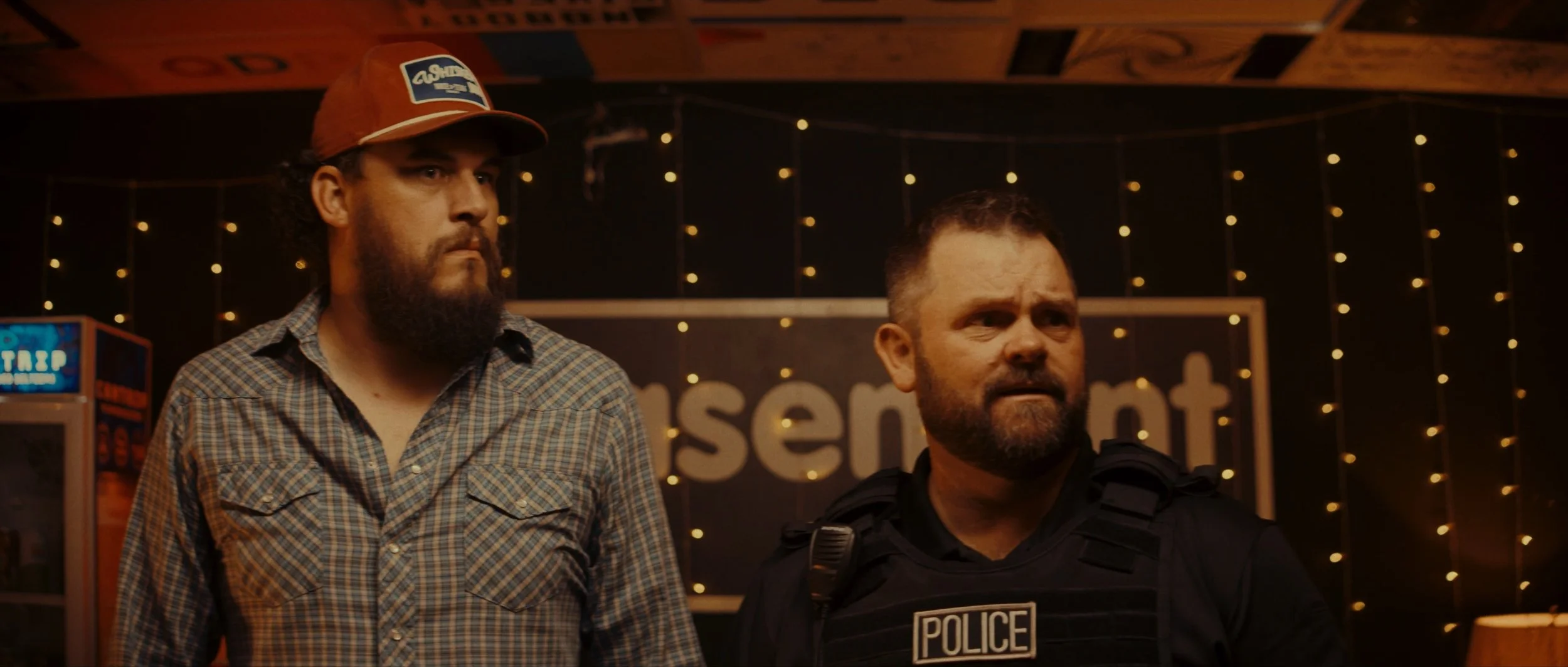Two men, one with a beard and wearing a cowboy hat, the other in a police uniform, standing inside a dimly lit room with small string lights and a sign that says 'presents' in the background.