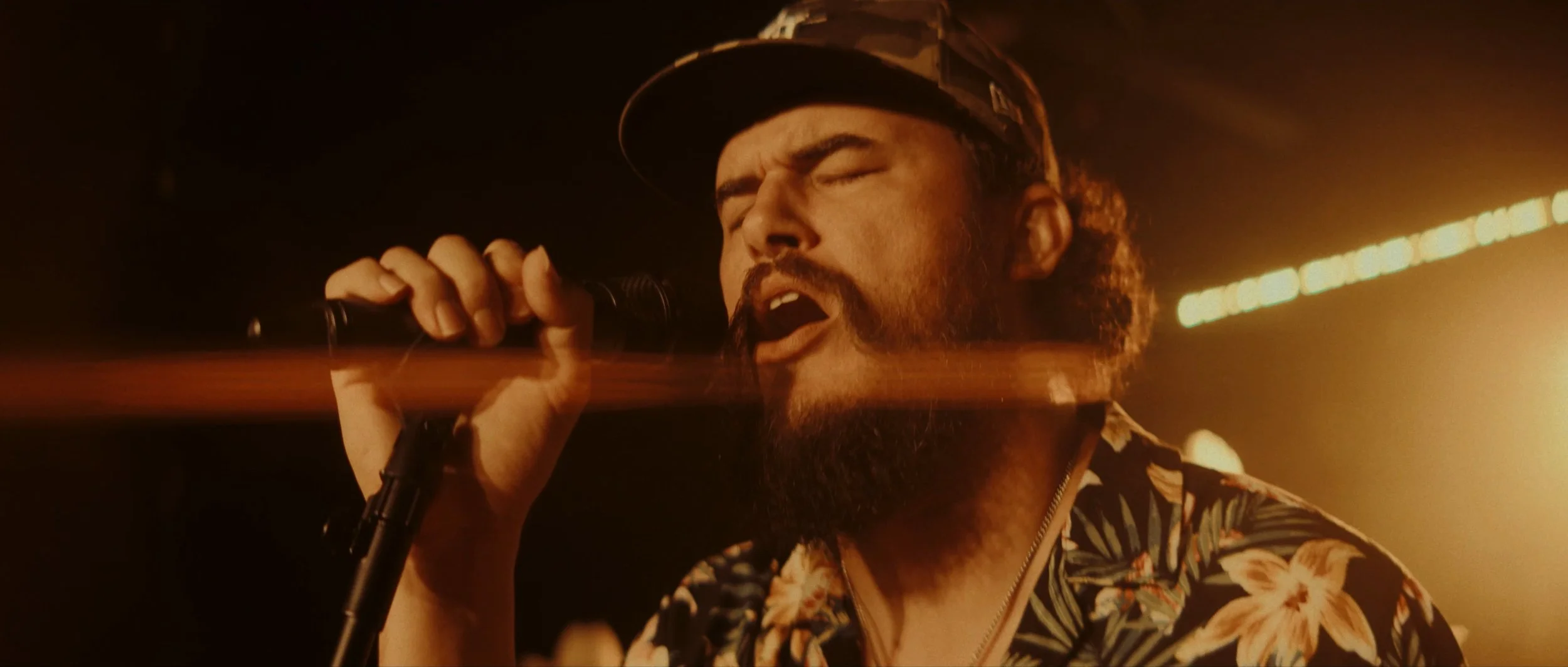 Man with a beard and mustache singing passionately into a microphone, wearing a patterned shirt and a cap, with a dark background and warm lighting.