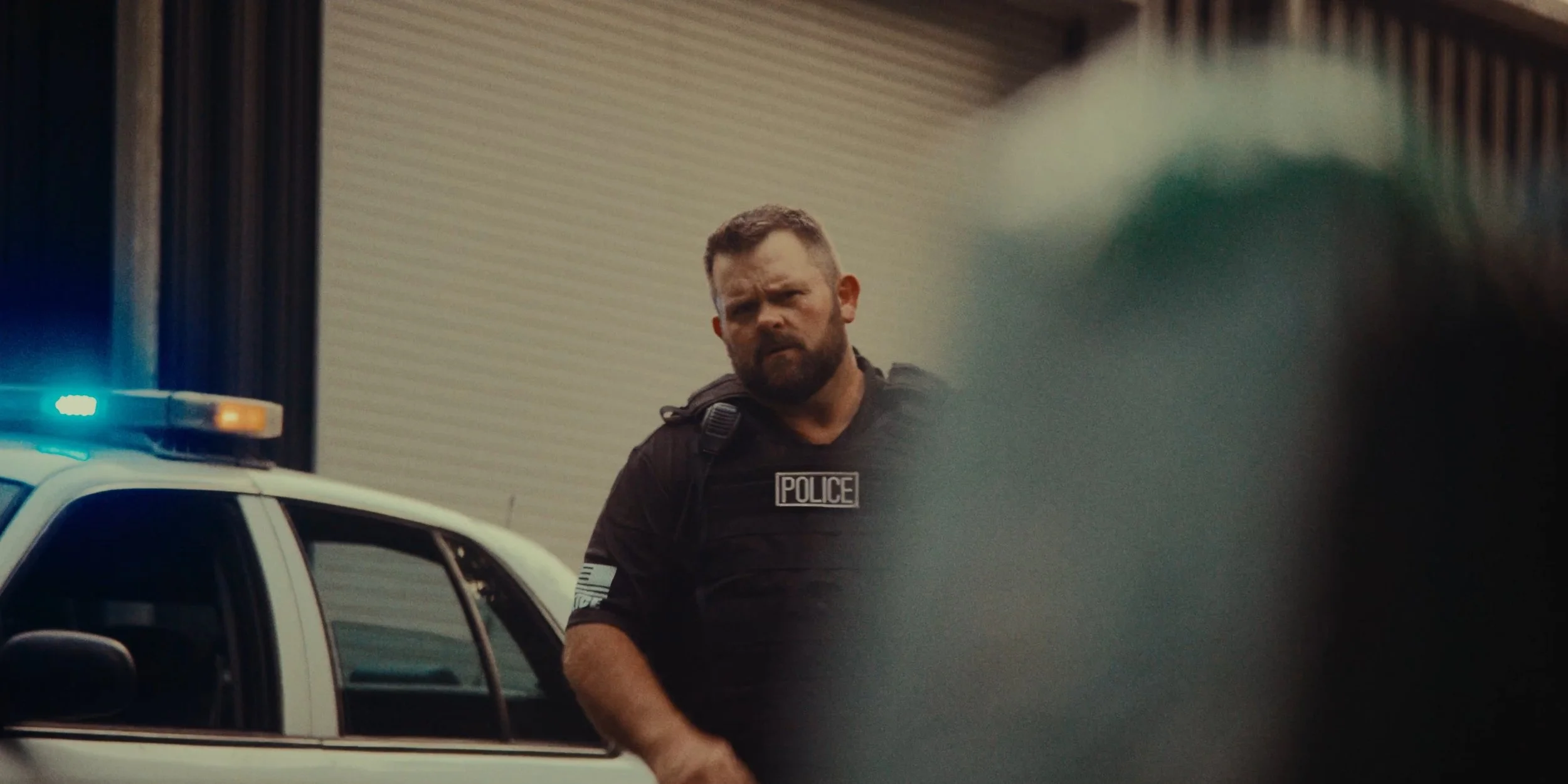 A police officer with a beard and short hair standing next to a police car with flashing blue and orange lights, looking to the right.