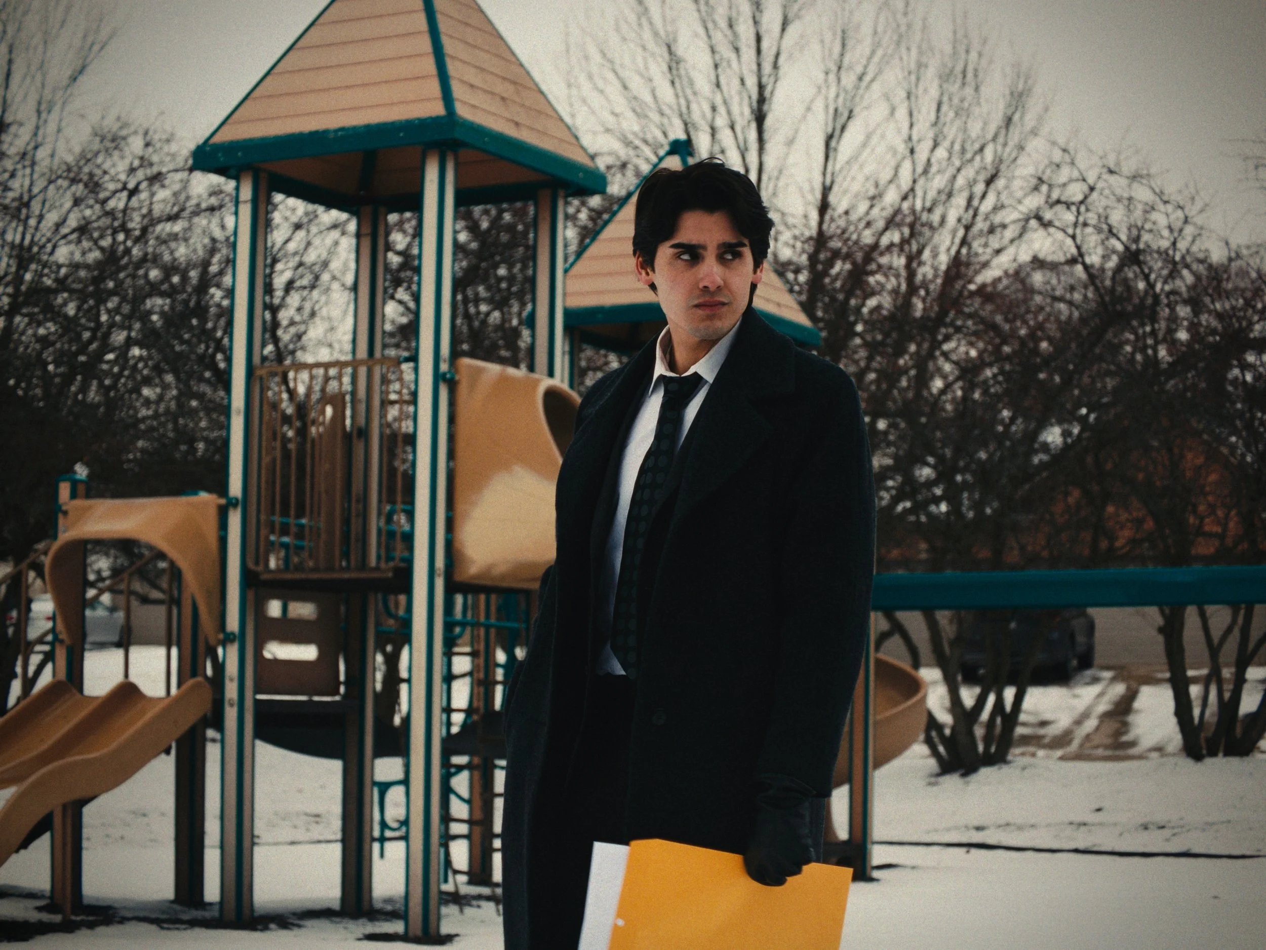 A young man in a black coat, white shirt, and tie stands outside a playground on a winter day, holding a yellow folder, with leafless trees in the background.