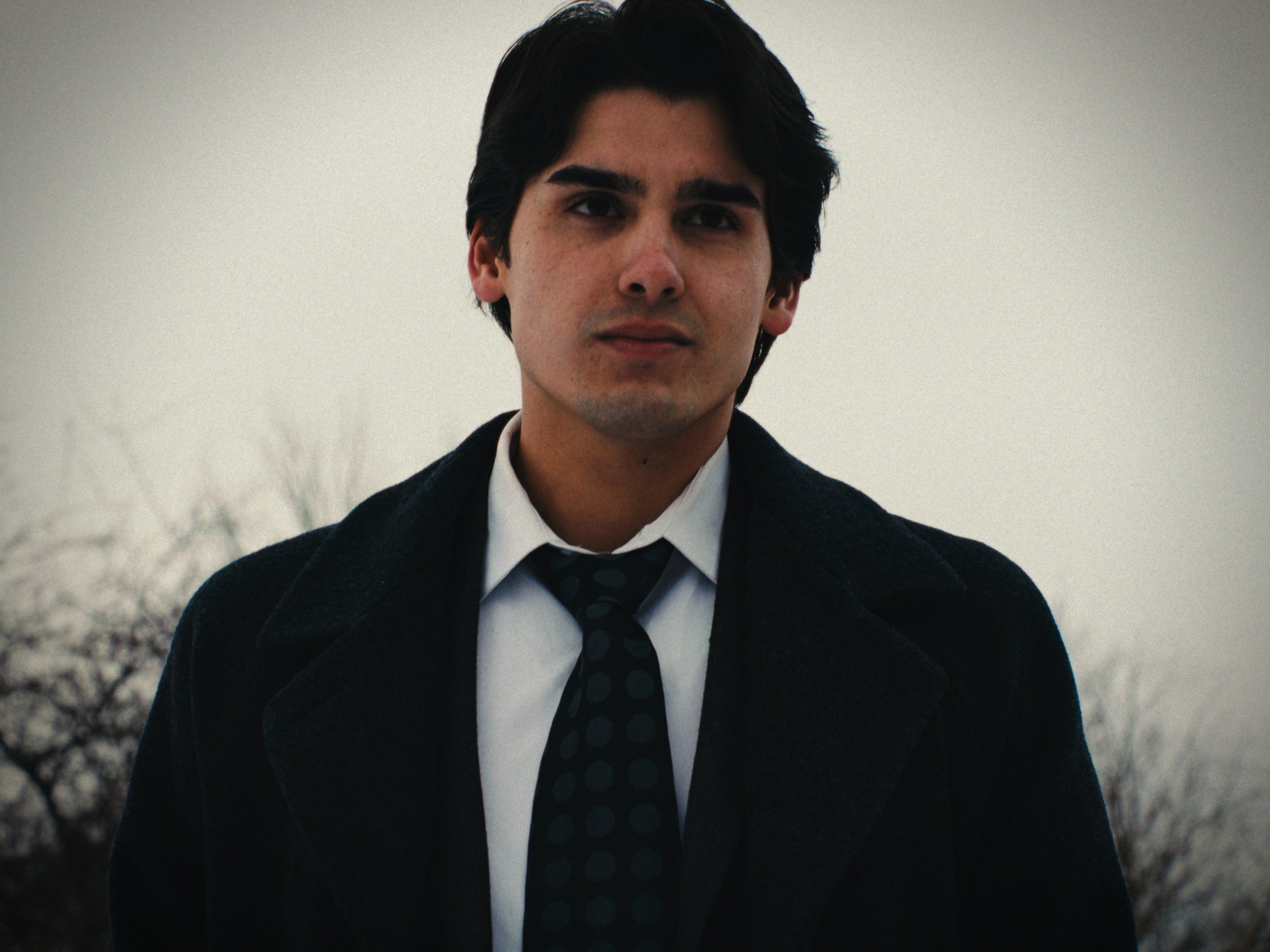 Young man with dark hair wearing a black coat, white shirt, and black dotted tie standing outdoors against a gray, overcast sky.