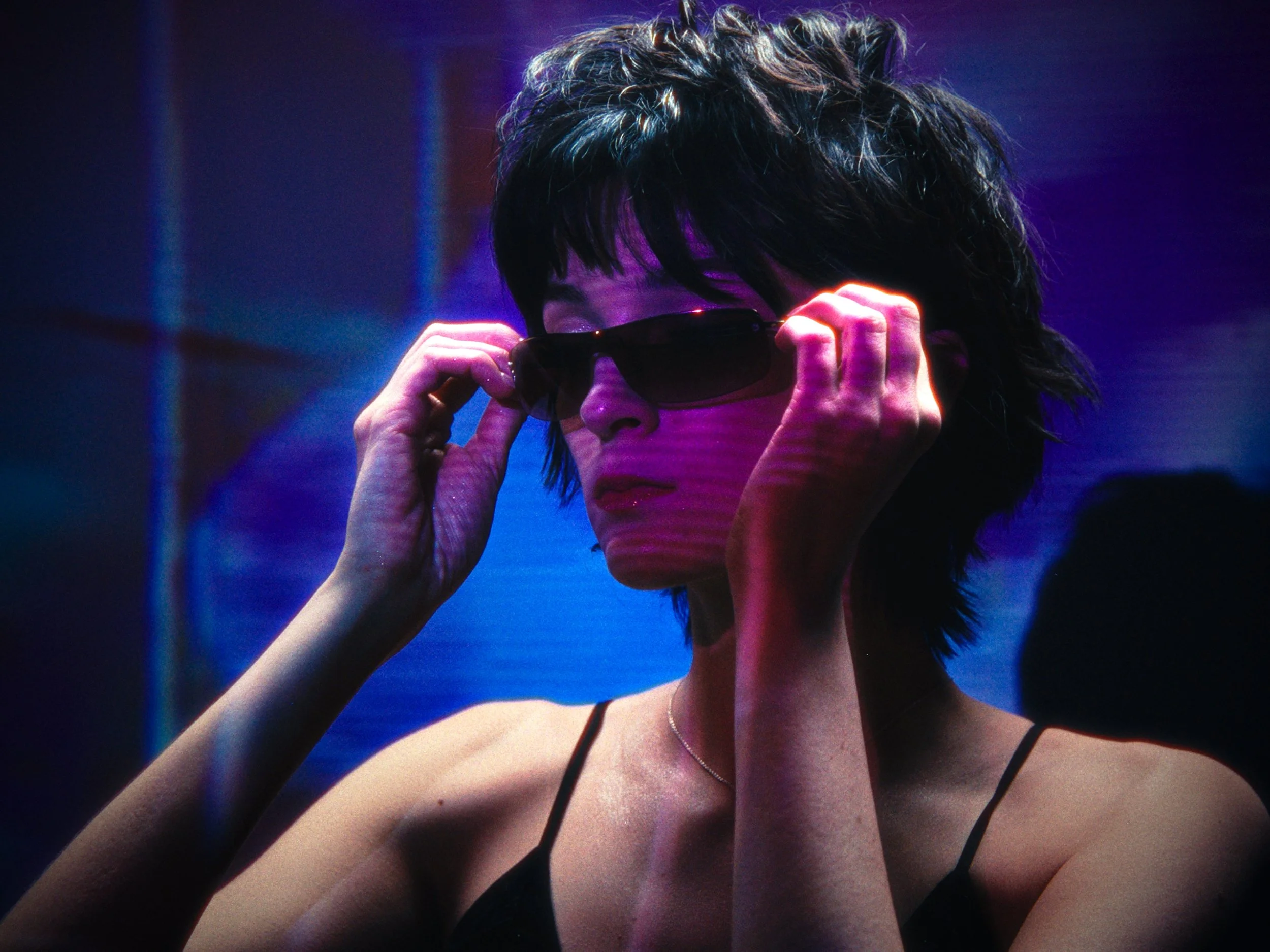 A woman with short dark hair and wearing sunglasses, holding the sunglasses with both hands, in a dimly lit environment with pink and blue neon lighting.