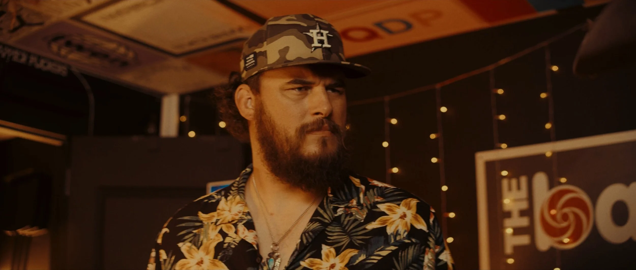 A man with a beard and mustache wearing a camouflage cap with a white logo, a floral Hawaiian shirt, and a necklace, inside a dimly lit venue with decorative string lights and a sign in the background.