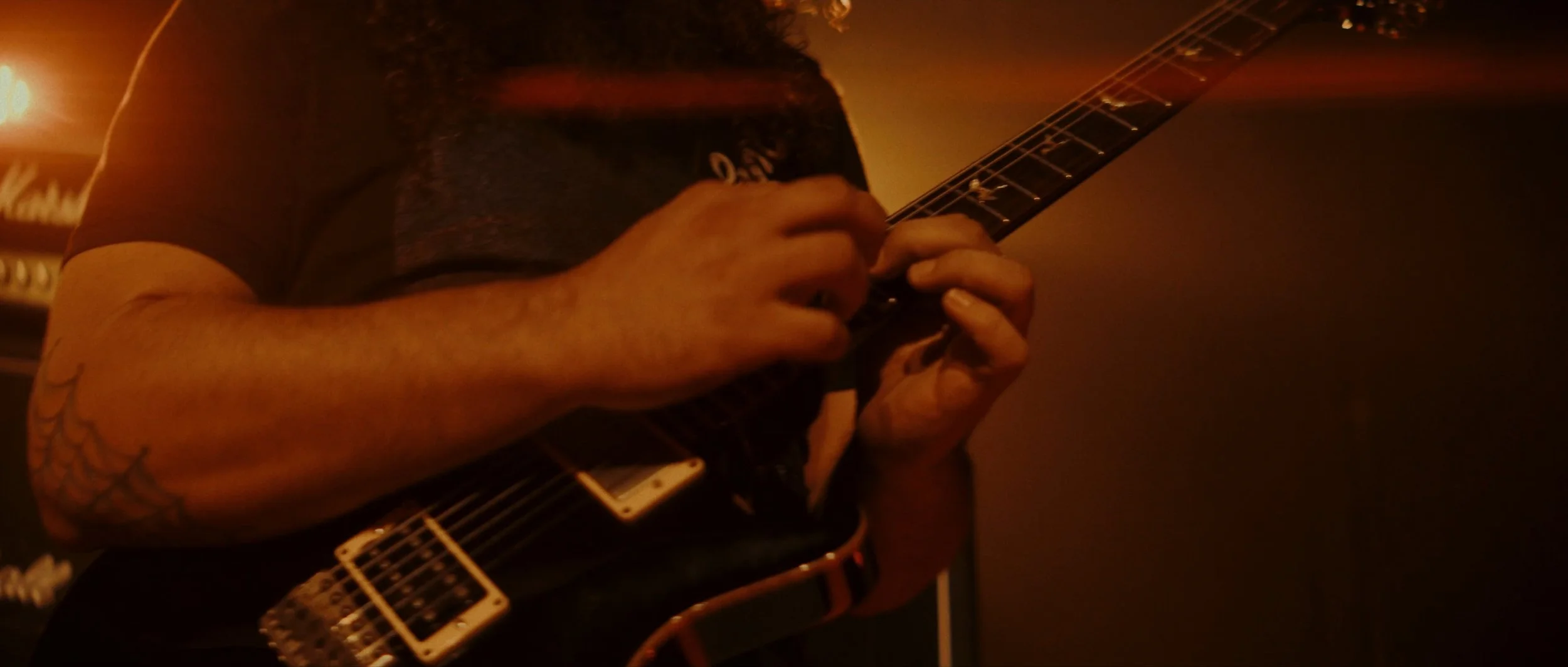 Person playing electric guitar in a dimly lit room with a warm orange glow, with amplifiers visible in the background.