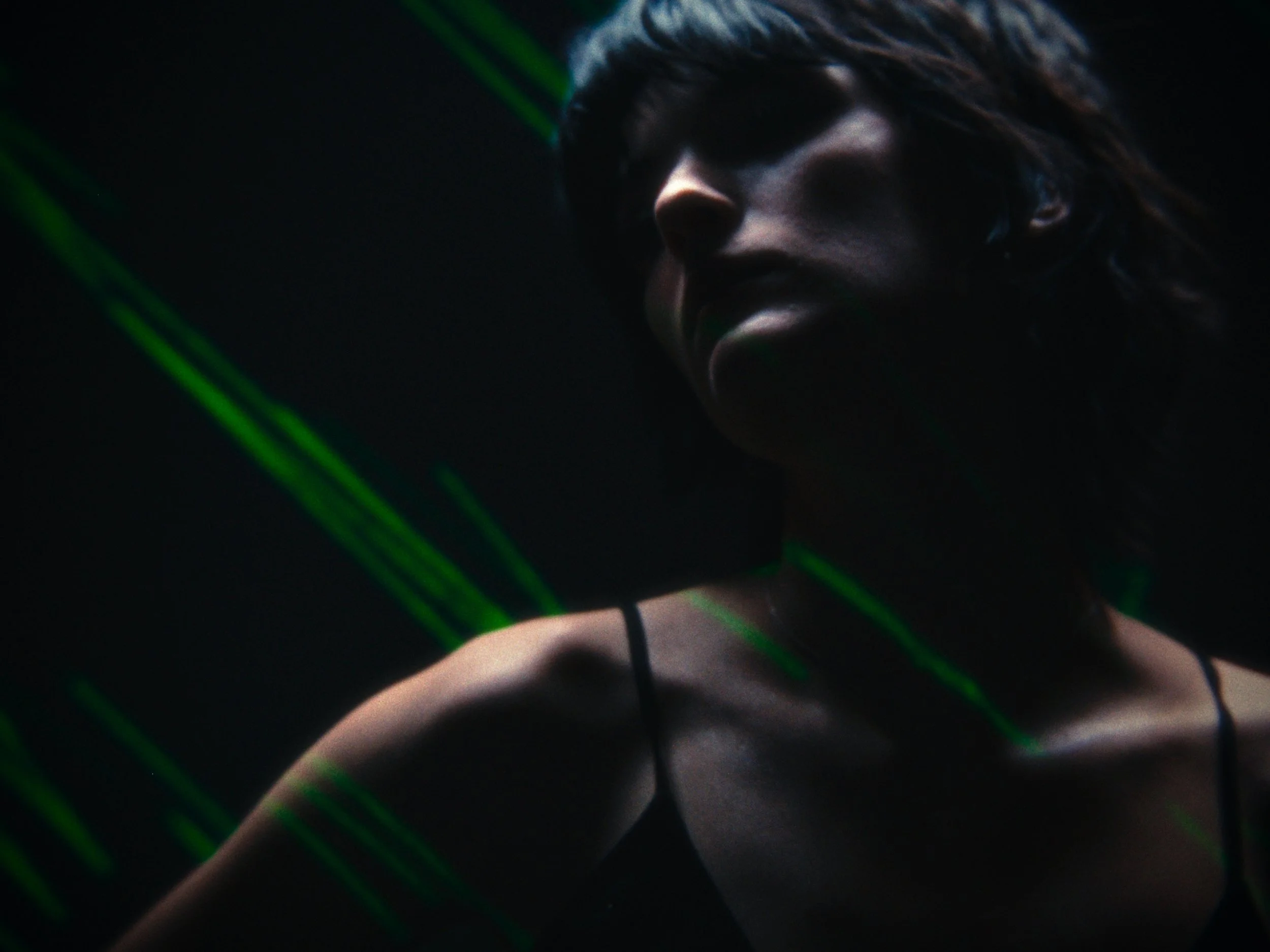 A woman in darkness with green laser beams crossing her face and shoulder, wearing a spaghetti strap top.