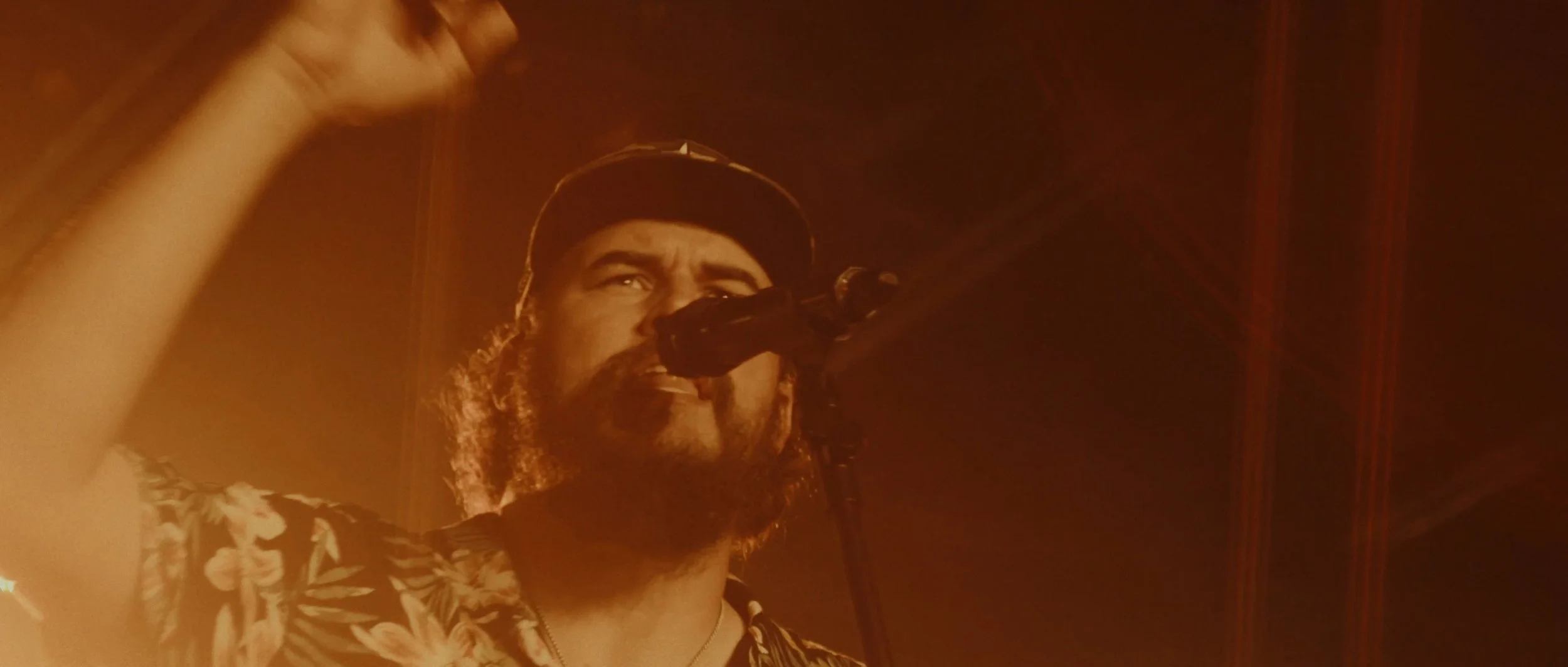 A man with a beard and curly hair, wearing a baseball cap and a Hawaiian shirt, singing into a microphone on stage with dim orange lighting.