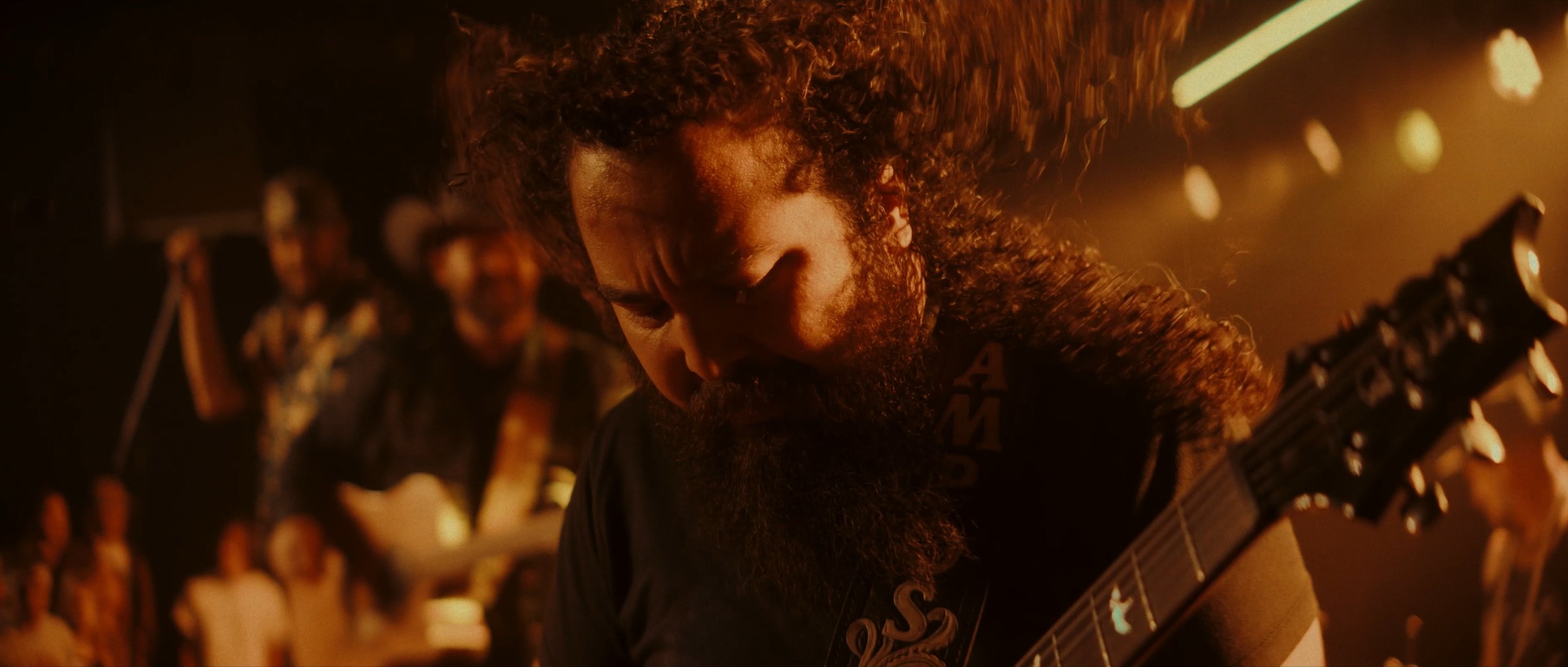 A bearded man with curly hair playing an electric guitar in a dimly lit room with an intense expression, with several people in the background.