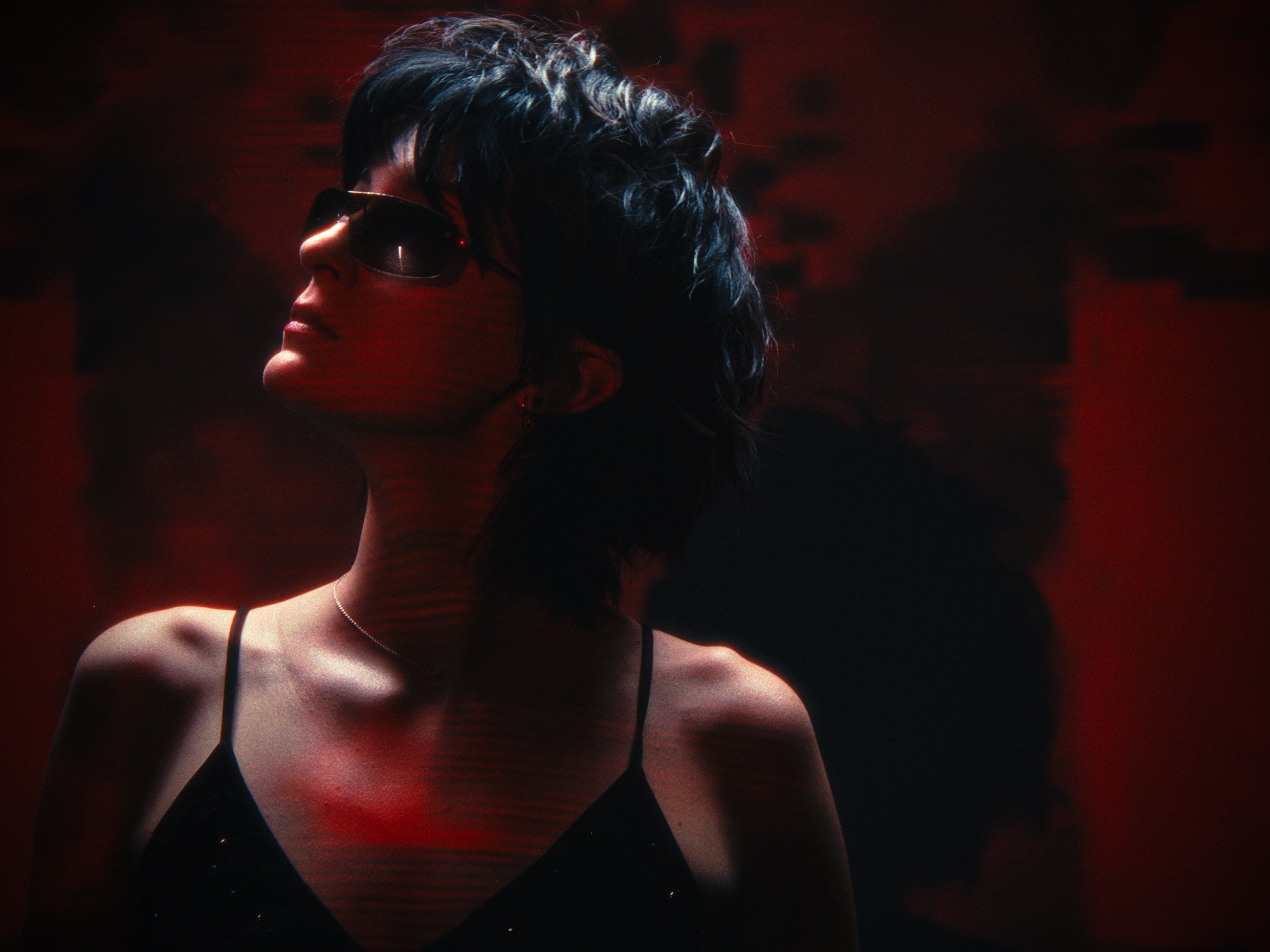 A woman with short dark hair wearing sunglasses and a black spaghetti strap top, standing against a dark, reddish background.
