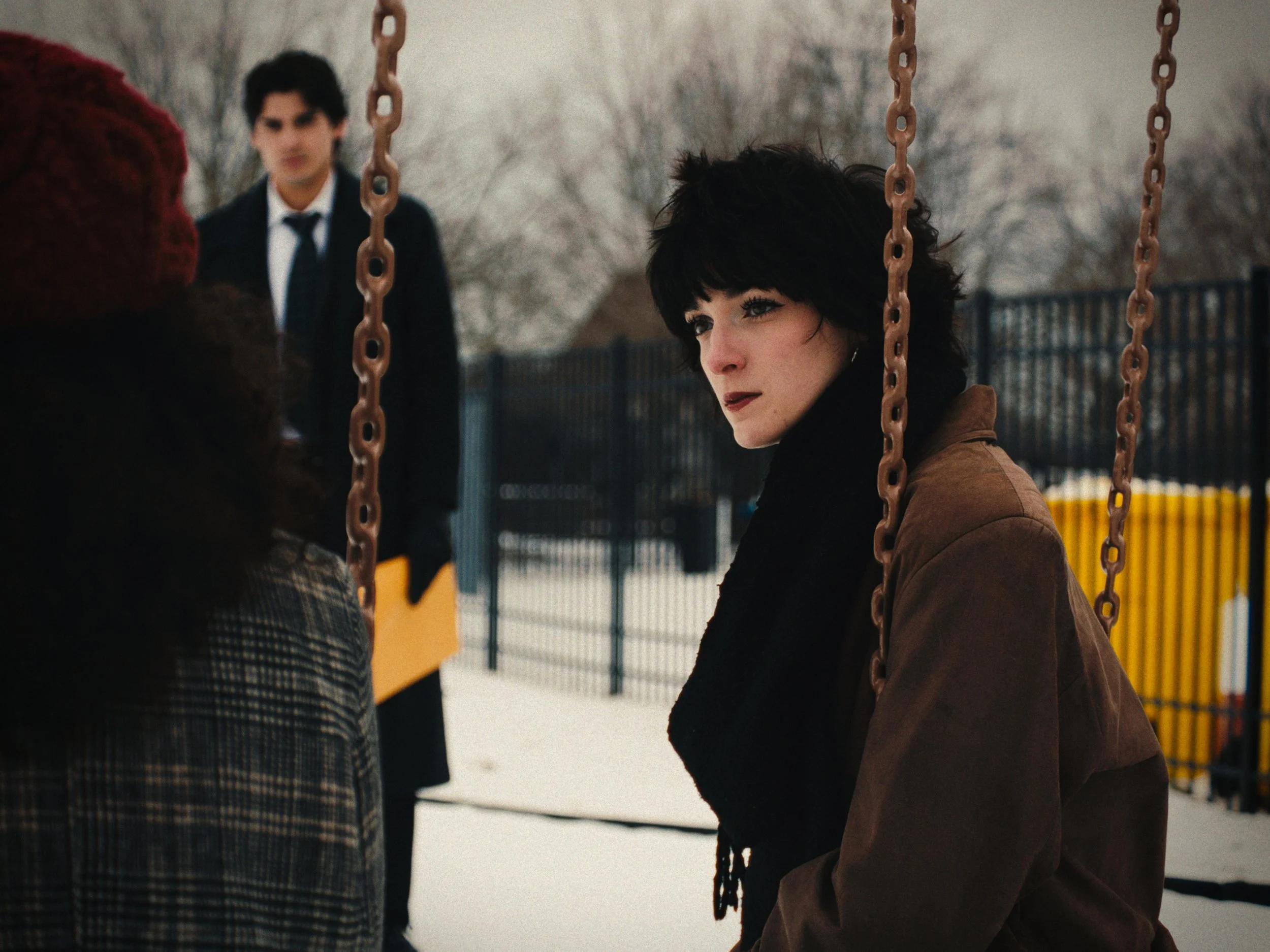 A woman with black hair and dark makeup sitting on a swing in a snowy park, looking at a person in front of her.