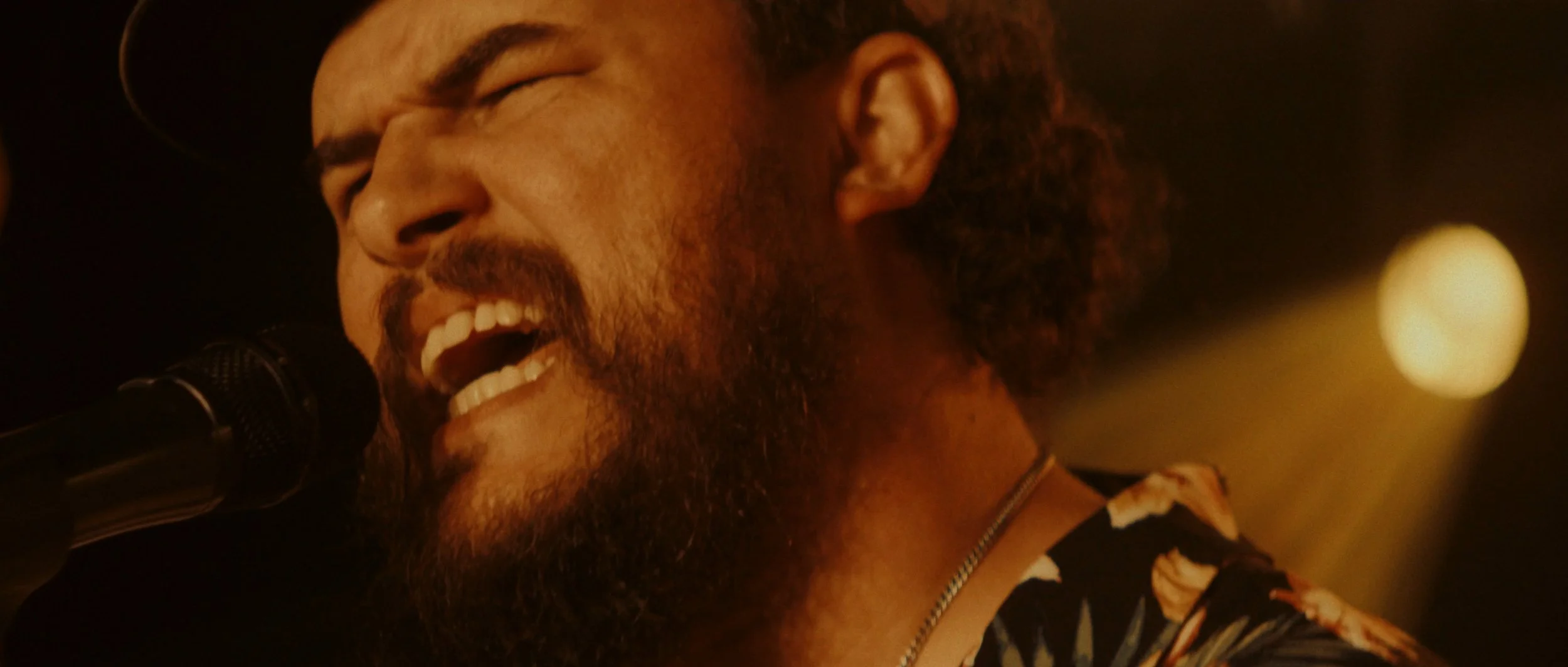 A man with curly hair and a beard singing passionately into a microphone, wearing a patterned shirt and a chain, with a warm, dimly lit background.