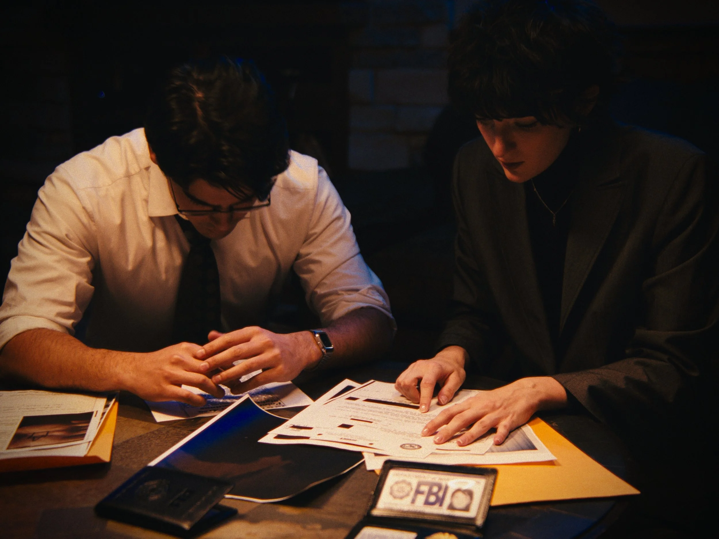 Two people, a man and a woman, sitting at a table reviewing documents and photographs, with some FBI related materials on the table.