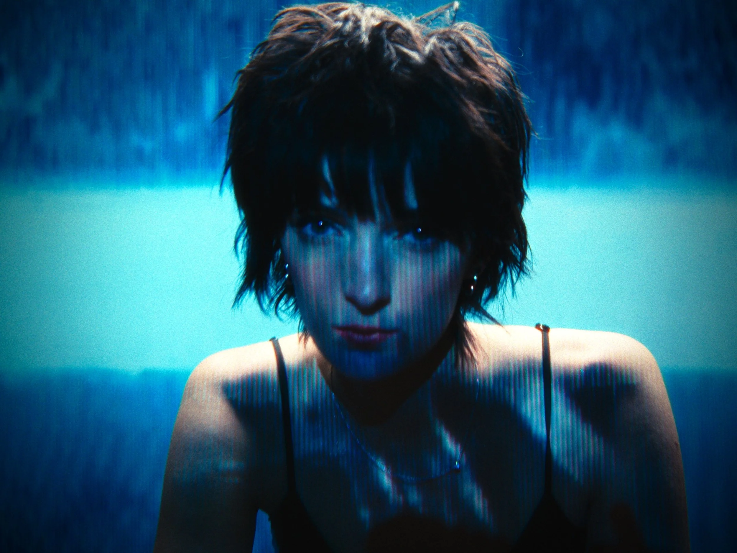 A woman with short, dark, shaggy hair, wearing a black spaghetti strap top, looking directly at the camera with a serious expression, against a blue background.