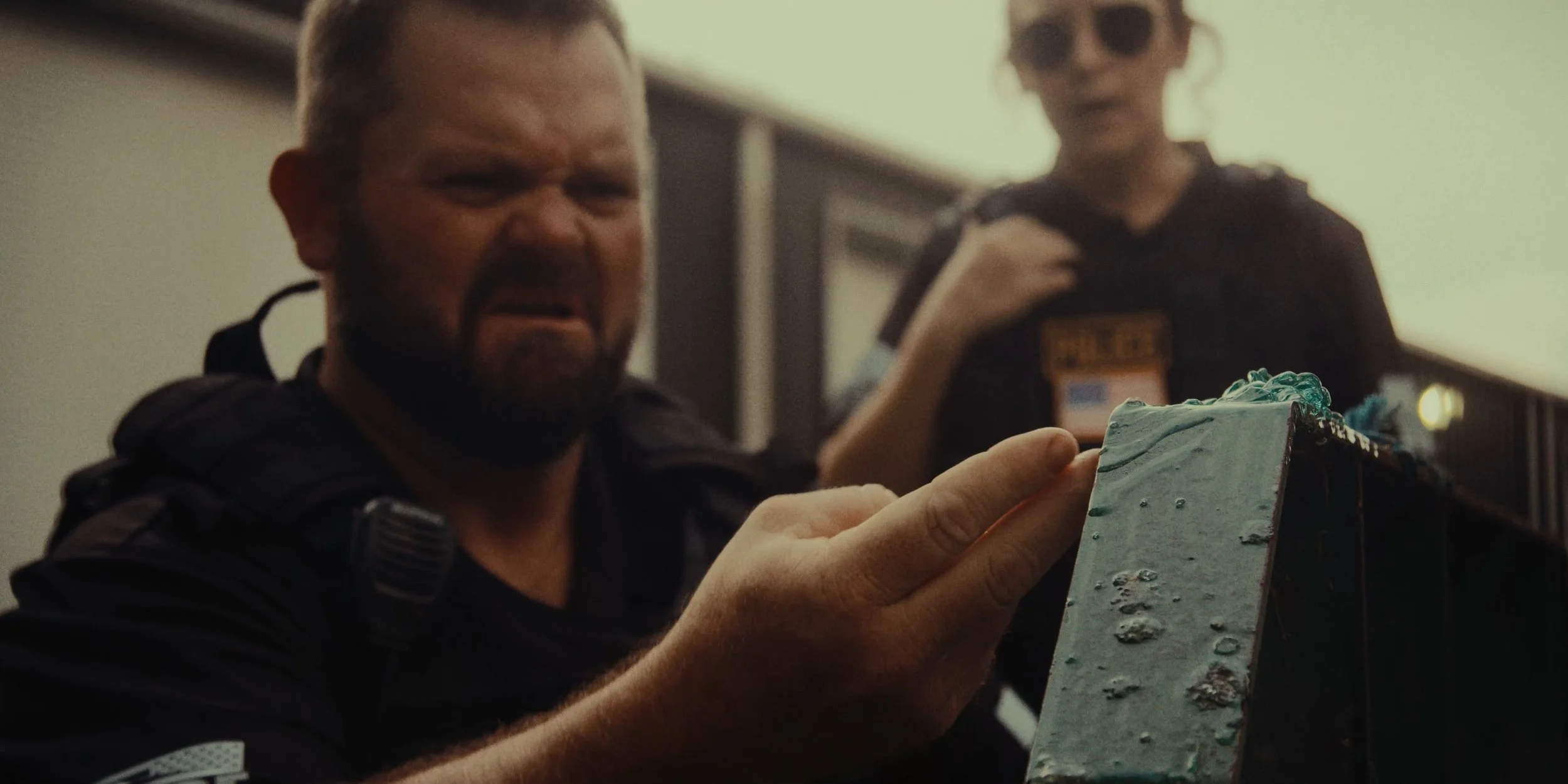 A man in a black uniform holding a piece of cake with a disgusted expression on his face, another person in the background wearing sunglasses and a badge.